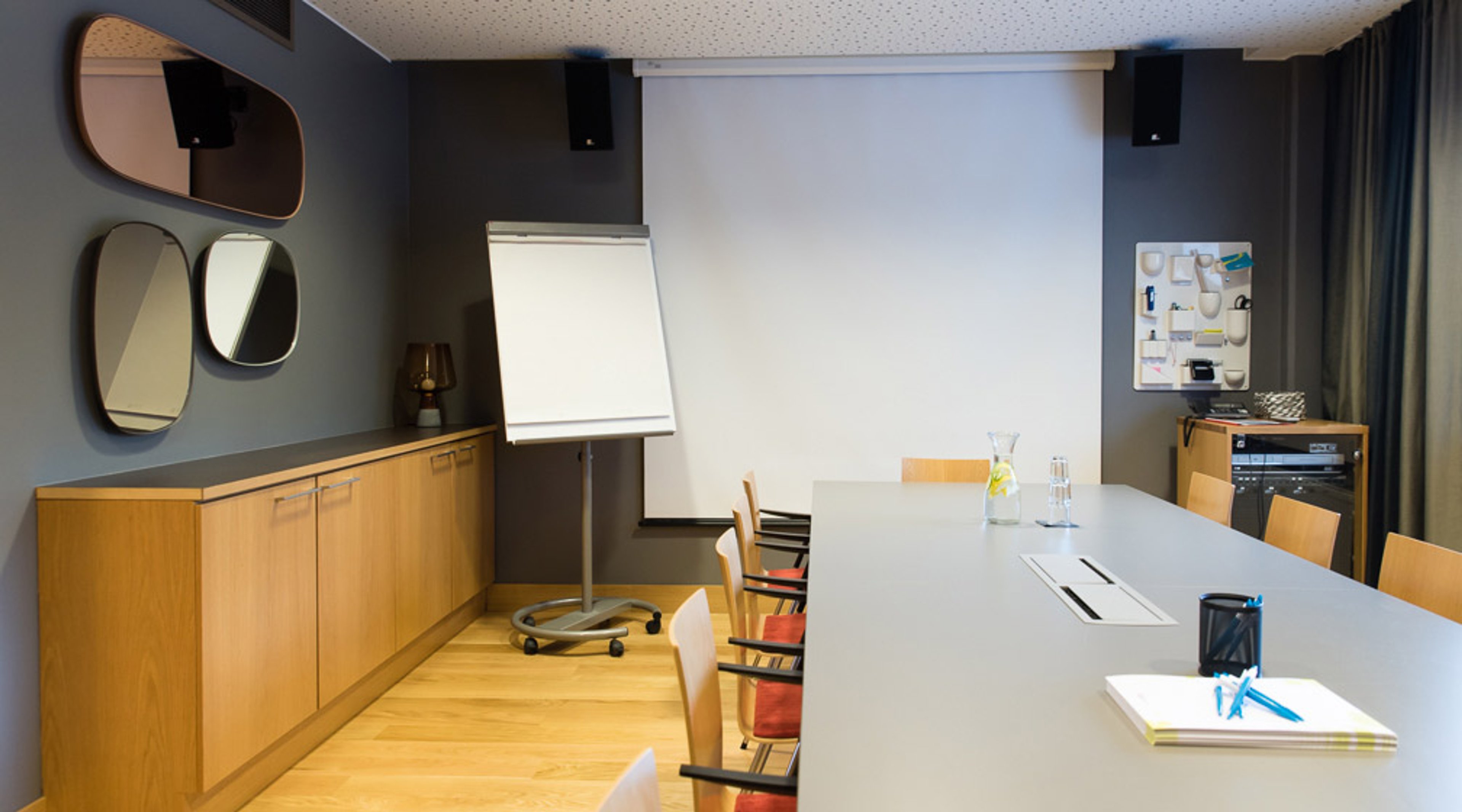 Moderni kokoushuone vaalealla puulattialla, harmaat seinät ja valkoinen pöytä. Flipchart-taulu ja projektiokangas edessä. Seinillä tyylikkäät peilit.
