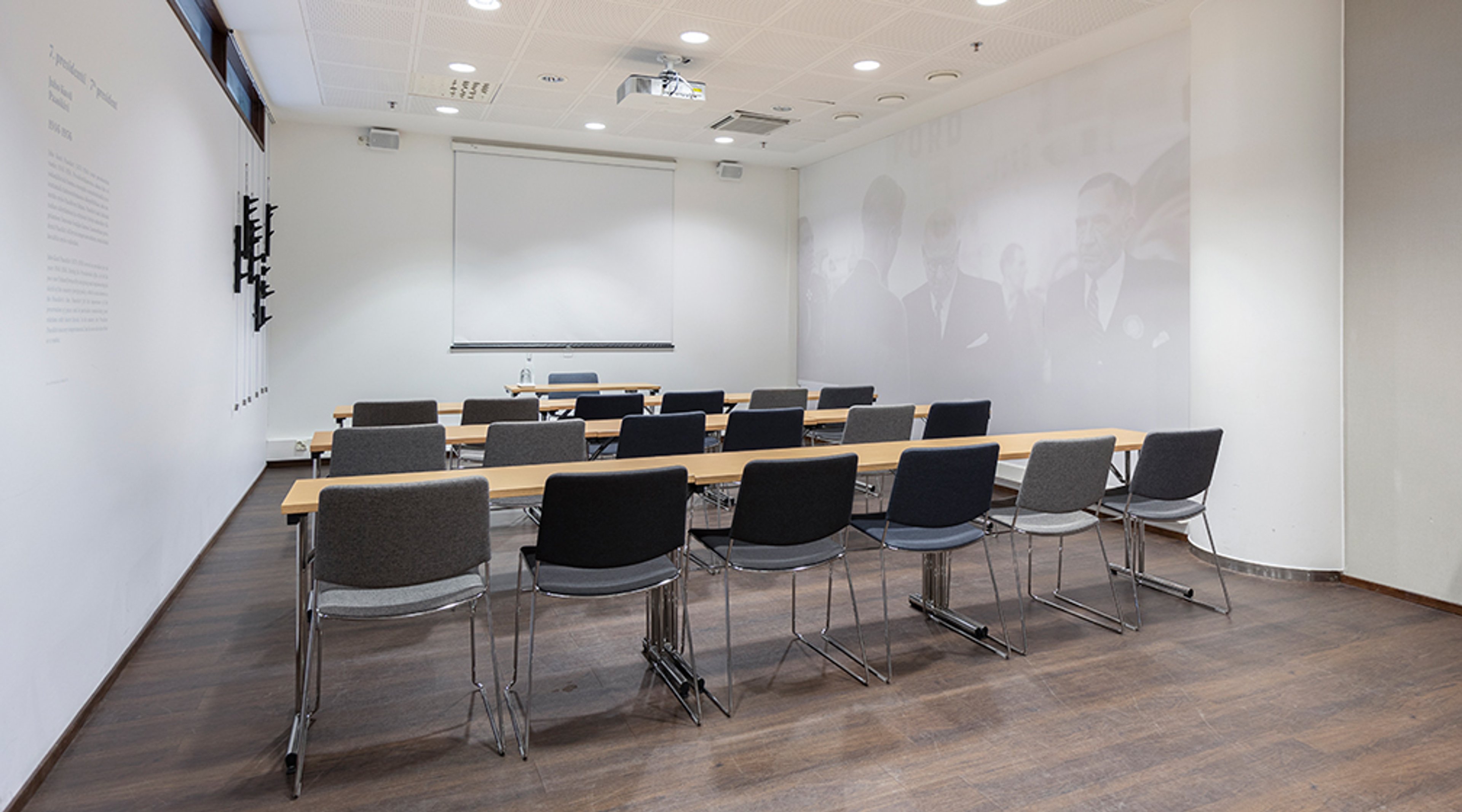 Pieni kokoustila, jossa puiset pöydät ja harmaat tuolit järjestettynä riveihin. Edessä valkoinen projektorikangas ja puhujanpöytä. Puulattia ja hyvä valaistus.
