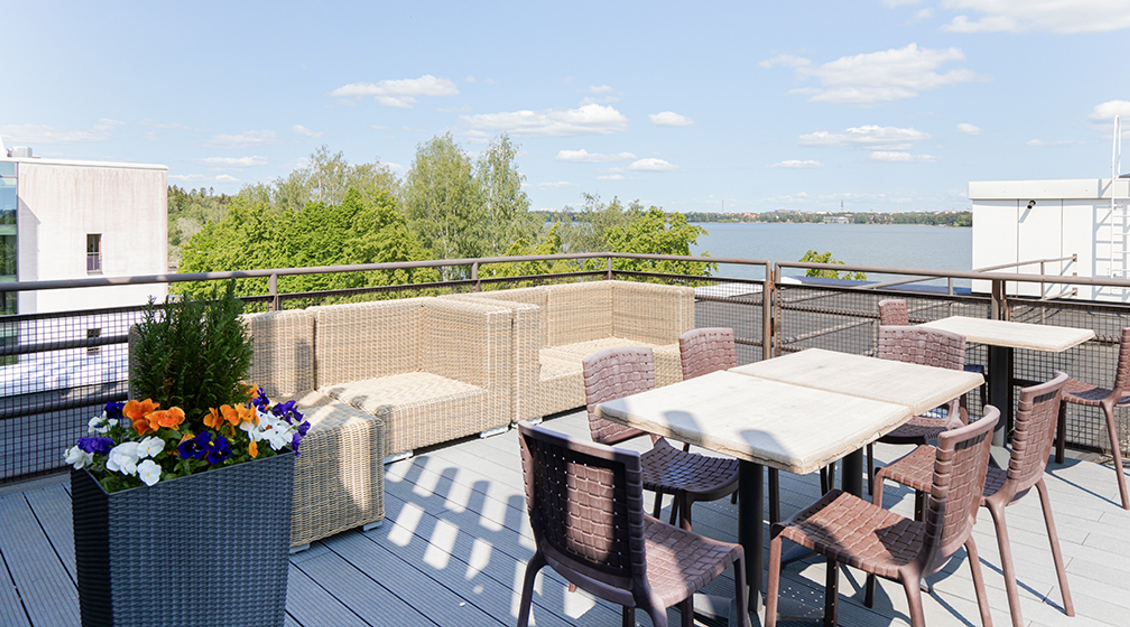 Kattoterassi järvinäkymällä, jossa rottinkikalusteita ja värikkäitä kukkia. Tasainen, esteetön lattia ja avara tila.