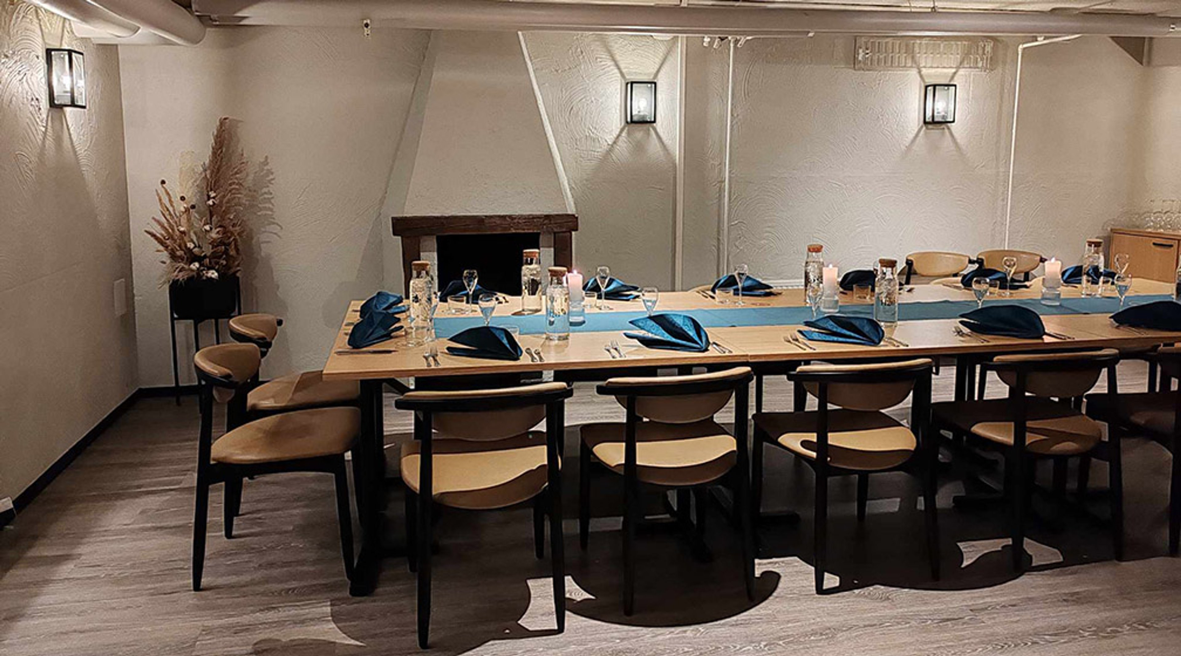 Kokoustila puisella pöydällä, sinisillä lautasliinoilla ja vesikarahveilla. Vaaleilla seinillä seinävalaisimet ja puulattia.