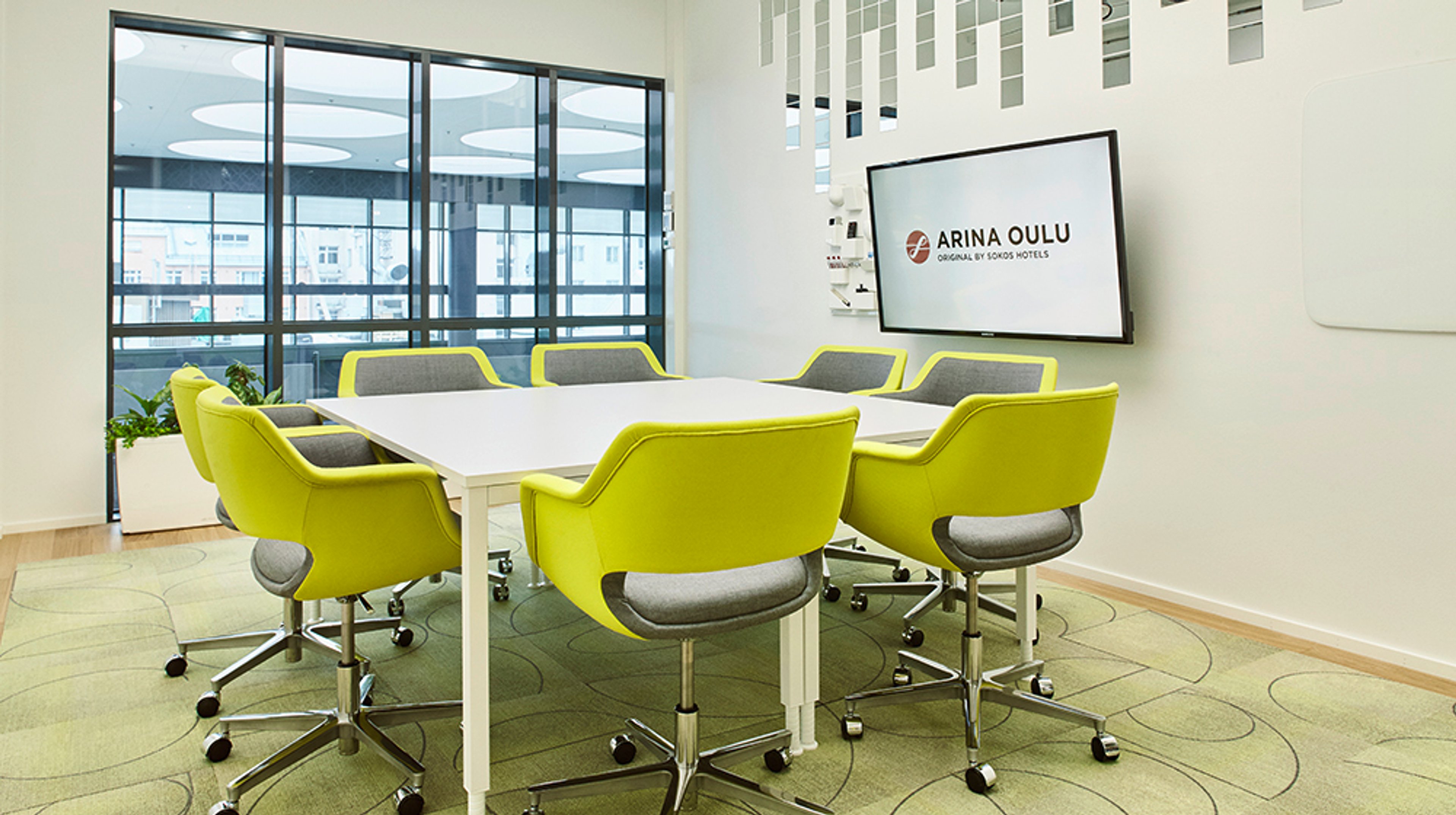 Moderni neuvottelutila, jossa on valkoinen pöytä, kirkkaan keltaiset tuolit, vihreä kokolattiamatto ja iso näyttö seinällä.