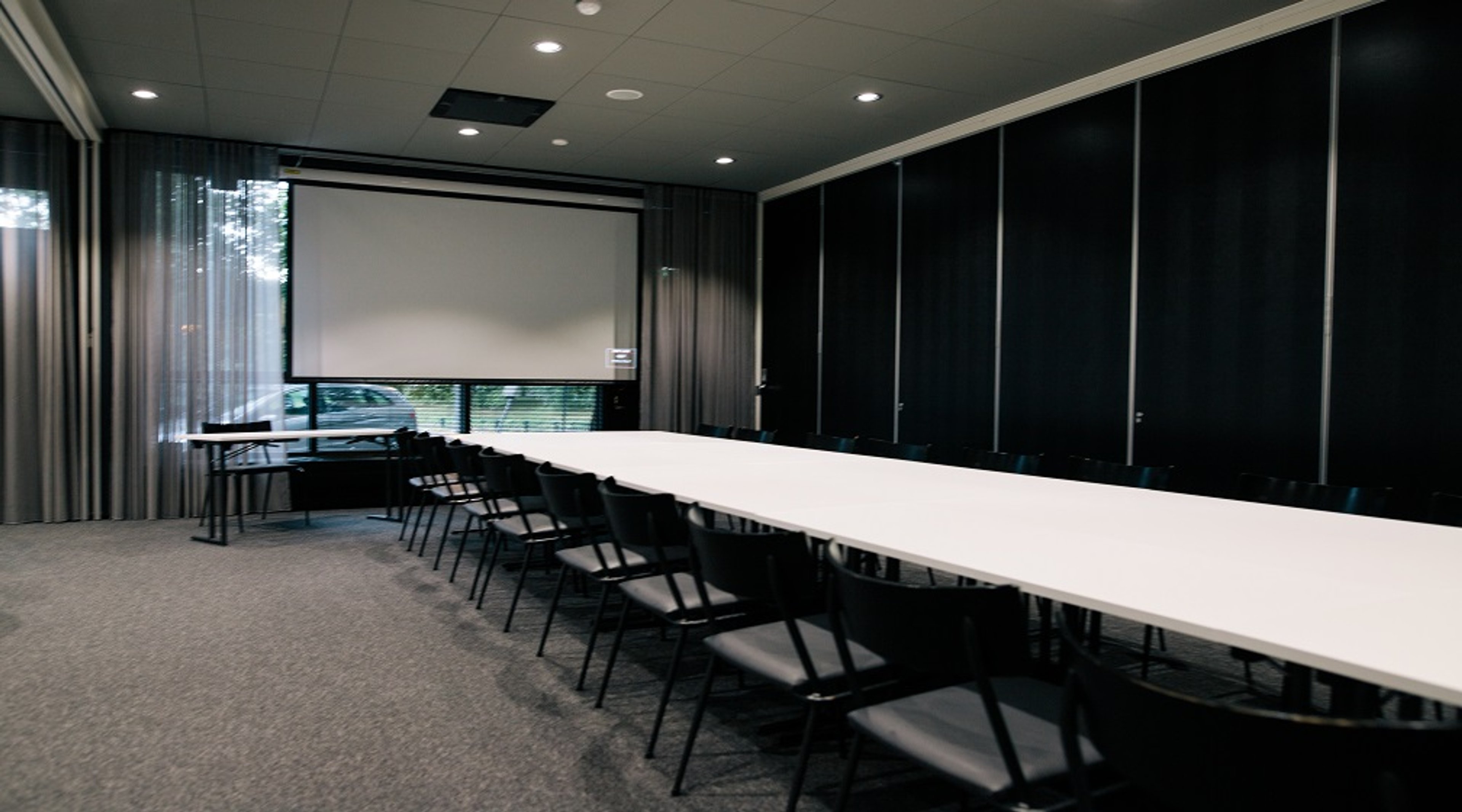 Kokoushuone pitkällä valkoisella pöydällä, mustilla tuoleilla ja valkoisella projektorikankaalla. Musta seinäpanelointi ja ikkunat.