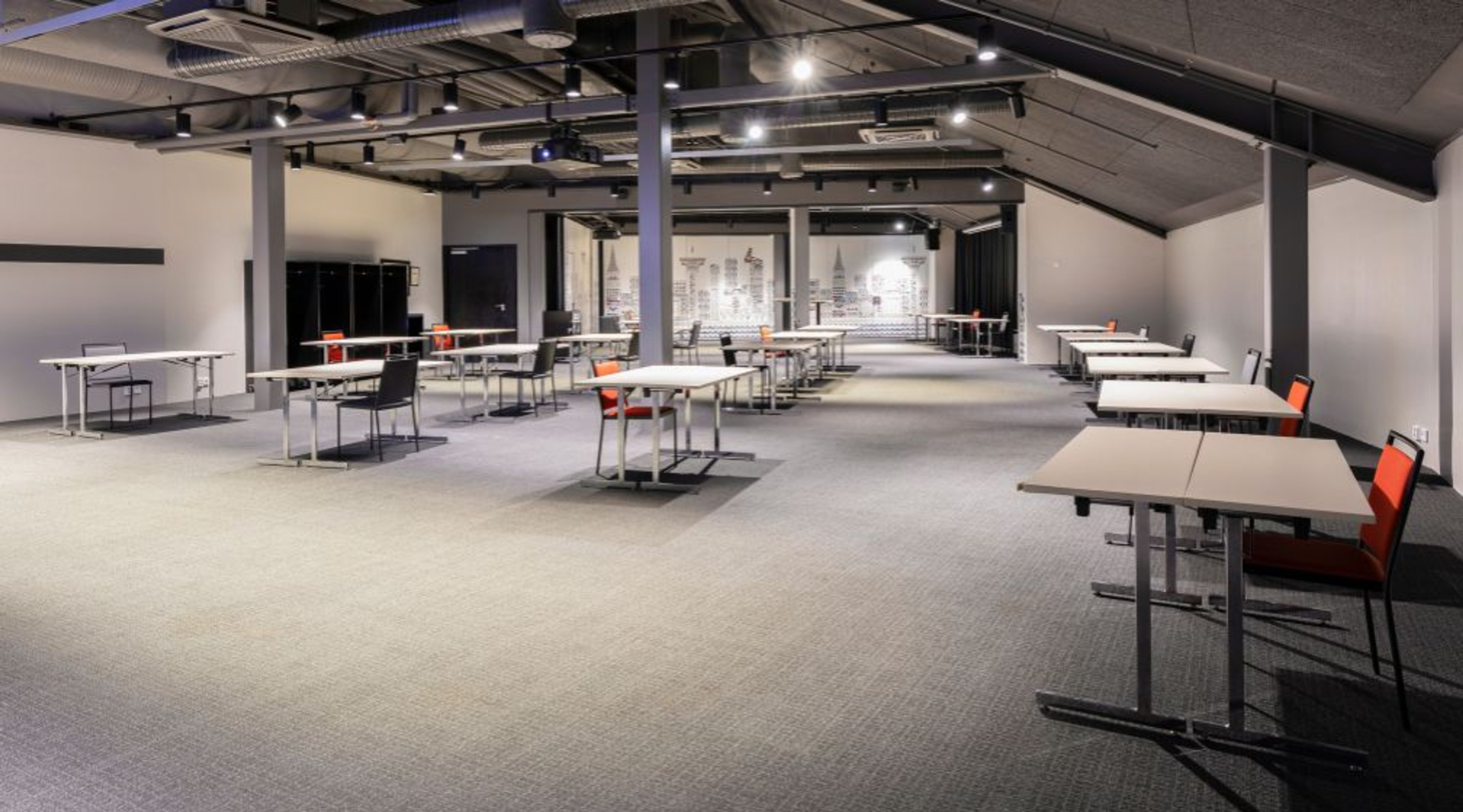 Avara konferenssi- tai työtila, jossa on tasainen mattolattiapinta, matala katto näkyvillä putkilla ja pilarein jäsennelty. Tilassa on hajallaan työpöytiä ja tuoleja.