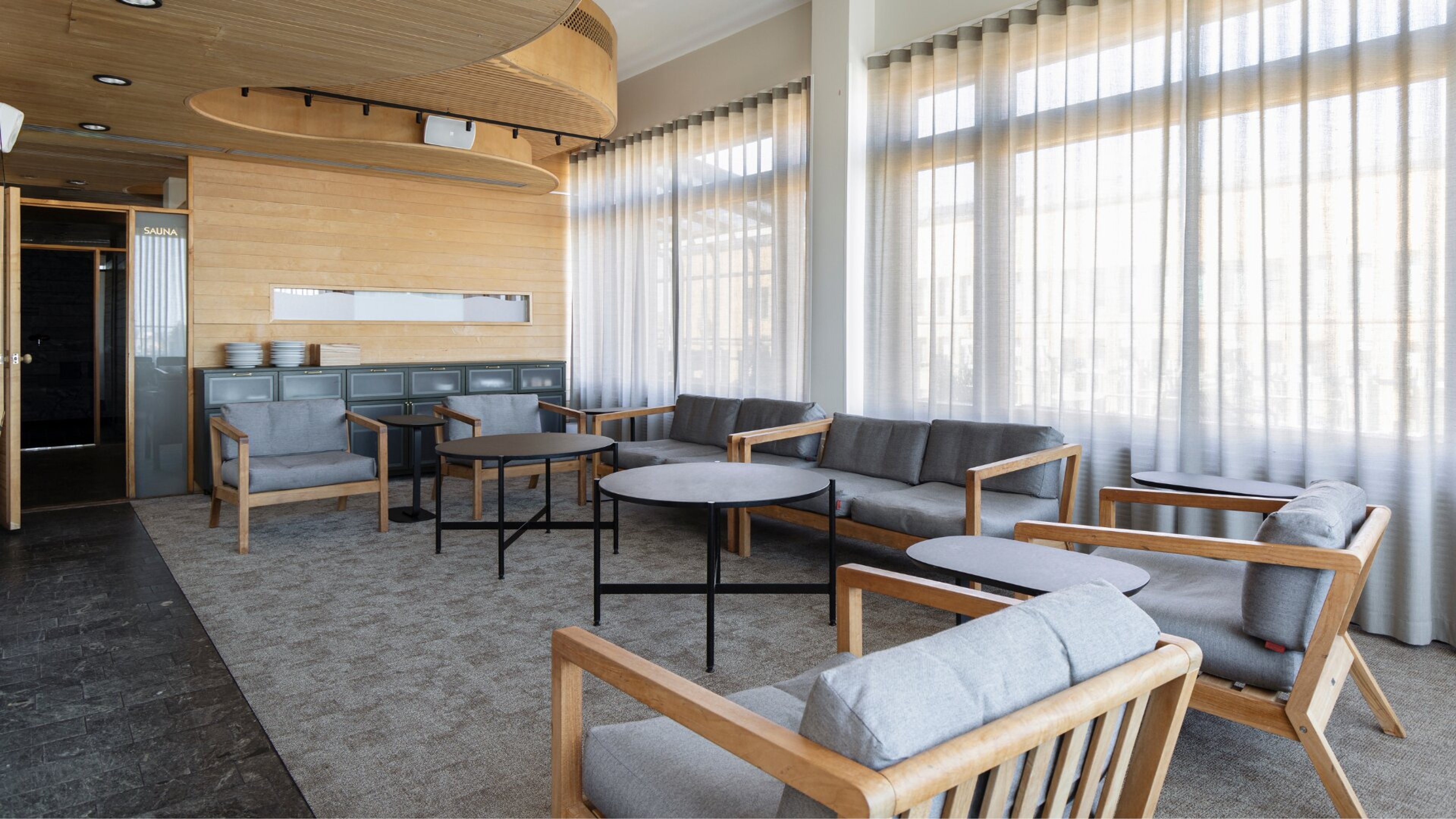 Saunatilan odotushuone, jossa harmaita sohvia ja nojatuoleja puukehyksillä, moderneja pöytiä ja vaaleat puuseinät. Isot ikkunat tuovat valoa.
