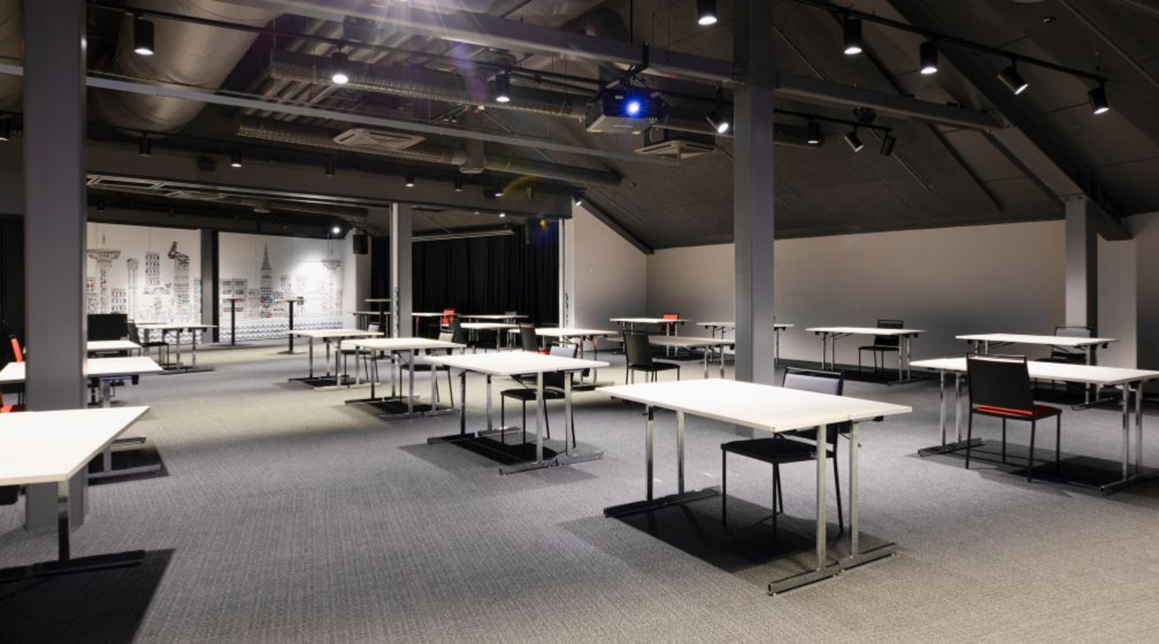 Moderni kokoustila, jossa pitkiä valkoisia pöytiä ja mustia tuoleja lyhytpinnoitteisella harmaalla moquette-lattialla. Korkea katto teollisella ilmastointikanavoinnilla ja hyvä valaistus.