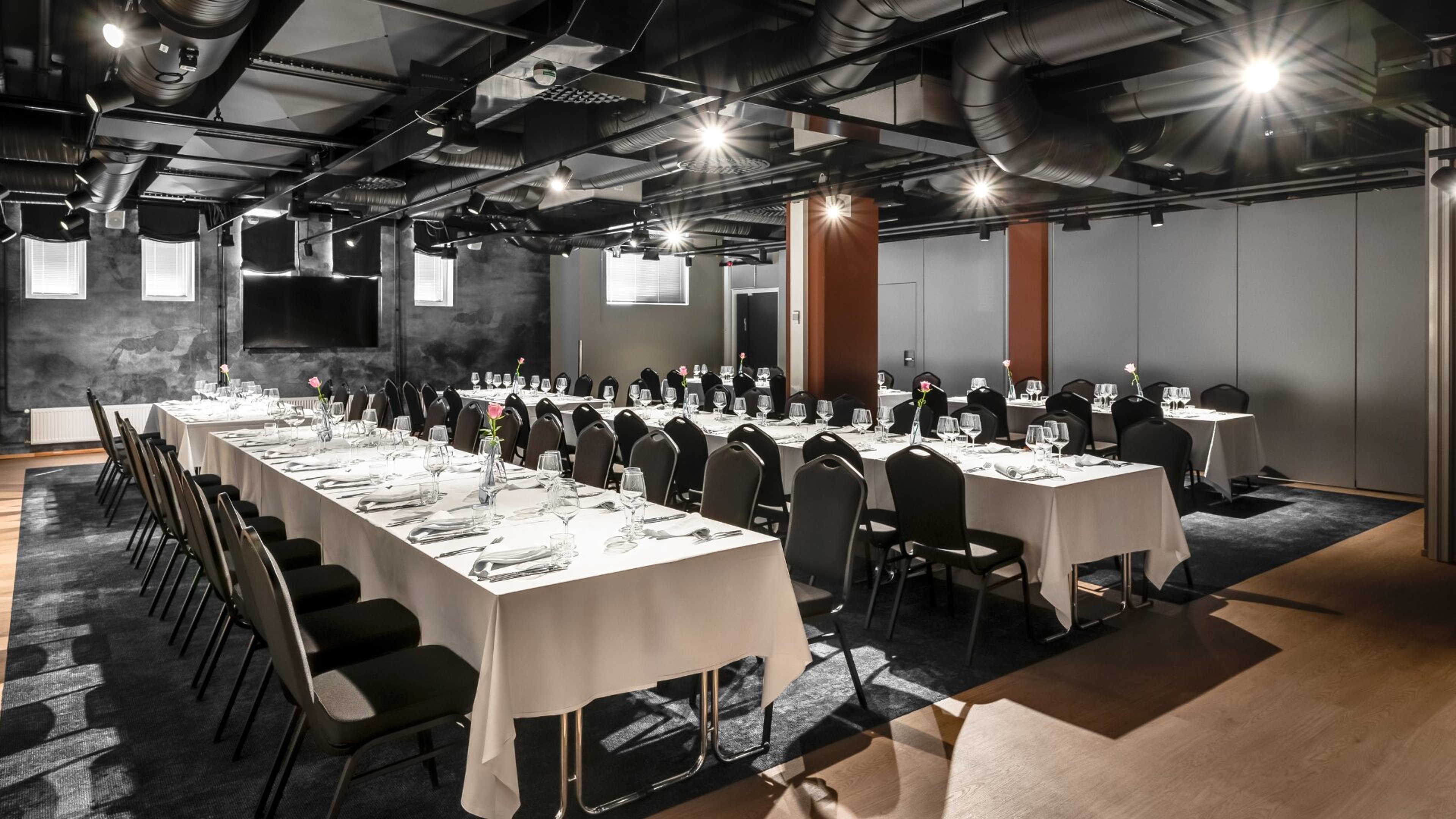 Moderni kokoustila jossa valkoliinaiset pöydät U-muodossa. Tummia tuoleja, lasiastiastoja ja pieniä kukkamaljakoita. Tummat seinät ja teollinen katto.