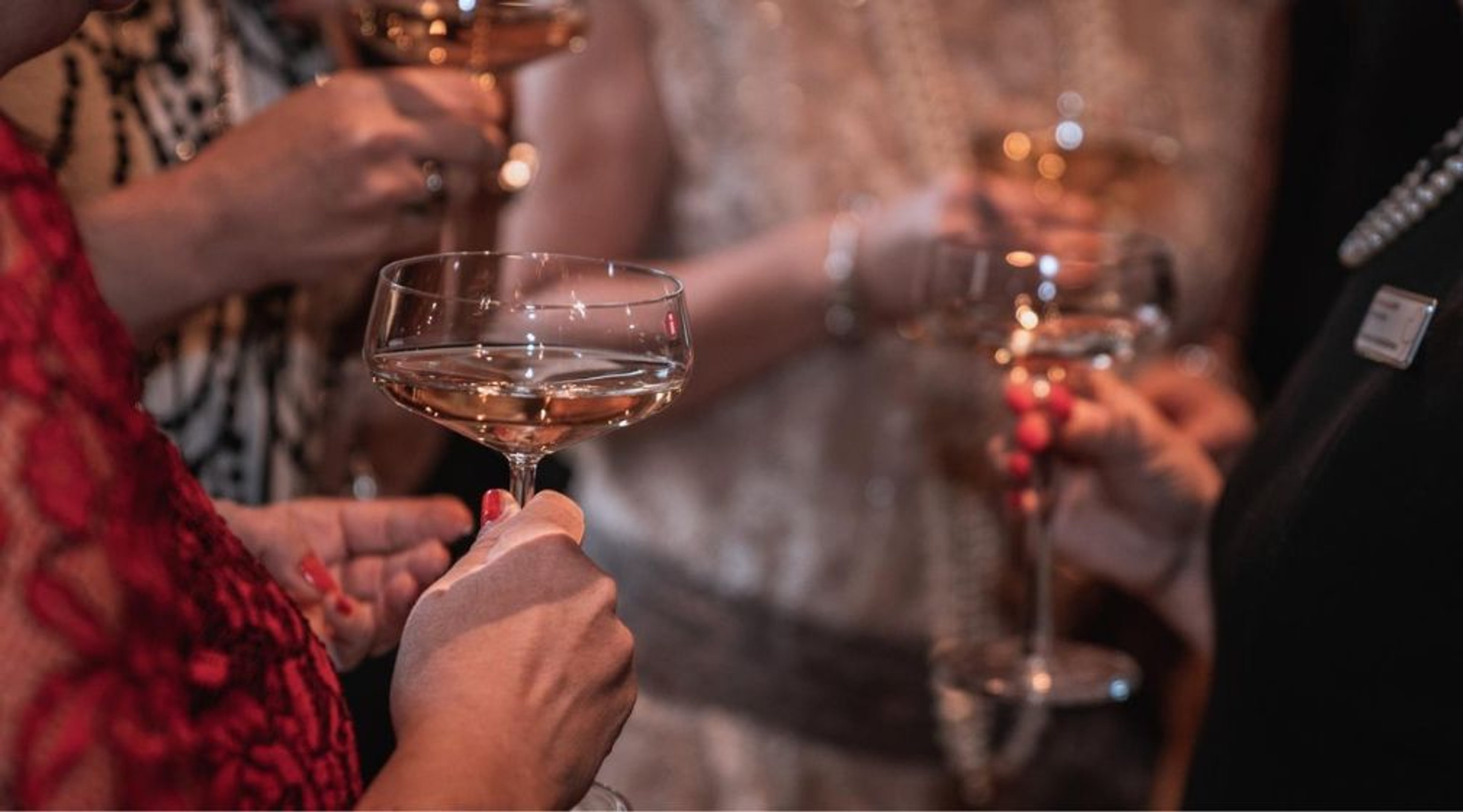 Person in red dress holding a champagne glass at an elegant gathering, with other guests with drinks visible in the softly lit background.