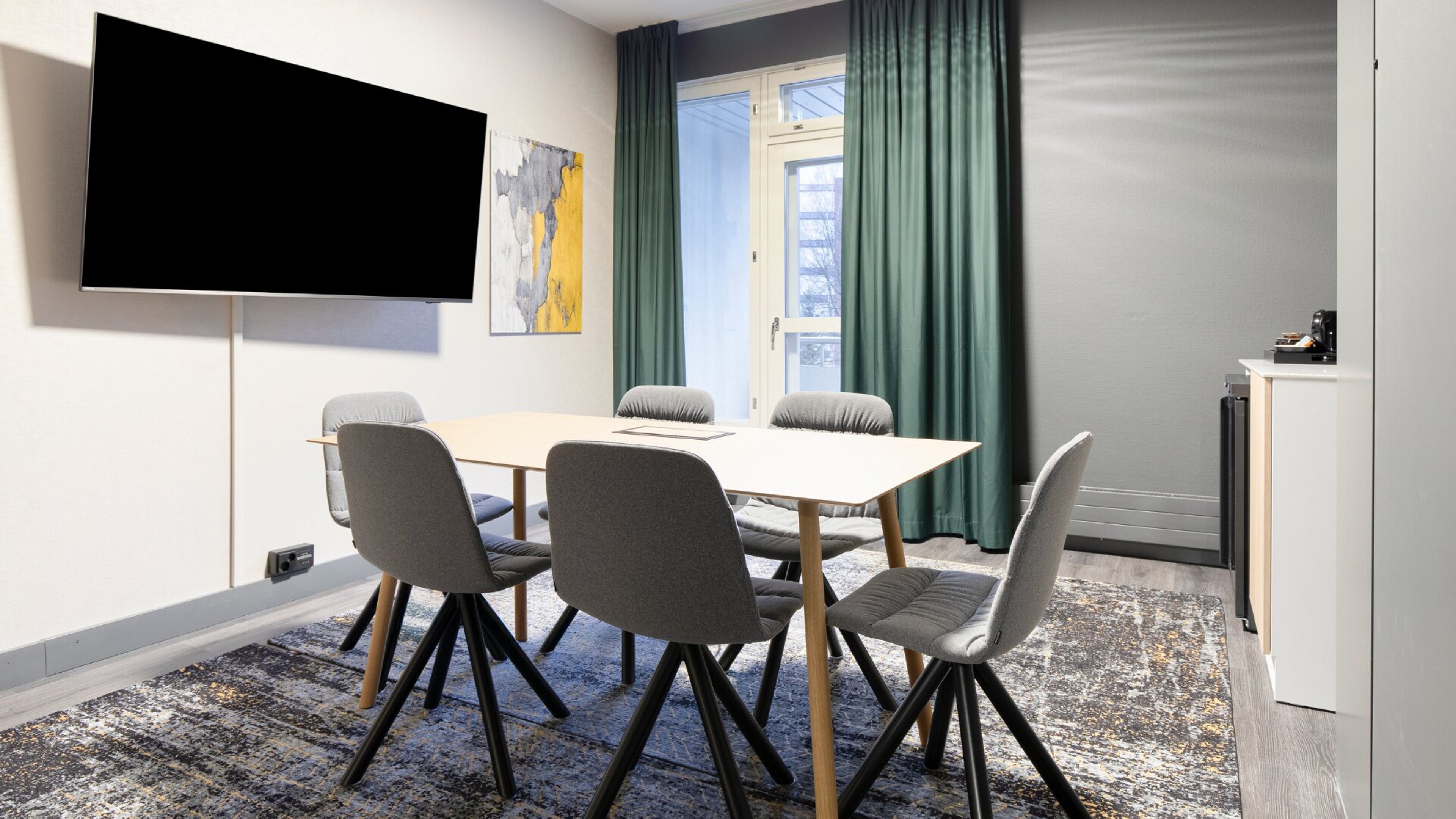 Moderni neuvotteluhuone, jossa puinen pöytä, kuusi harmaata tuolia, seinälle asennettu iso televisio ja vihreät verhot. Lattia on kova, ikkunasta näkyy luonnonvaloa.