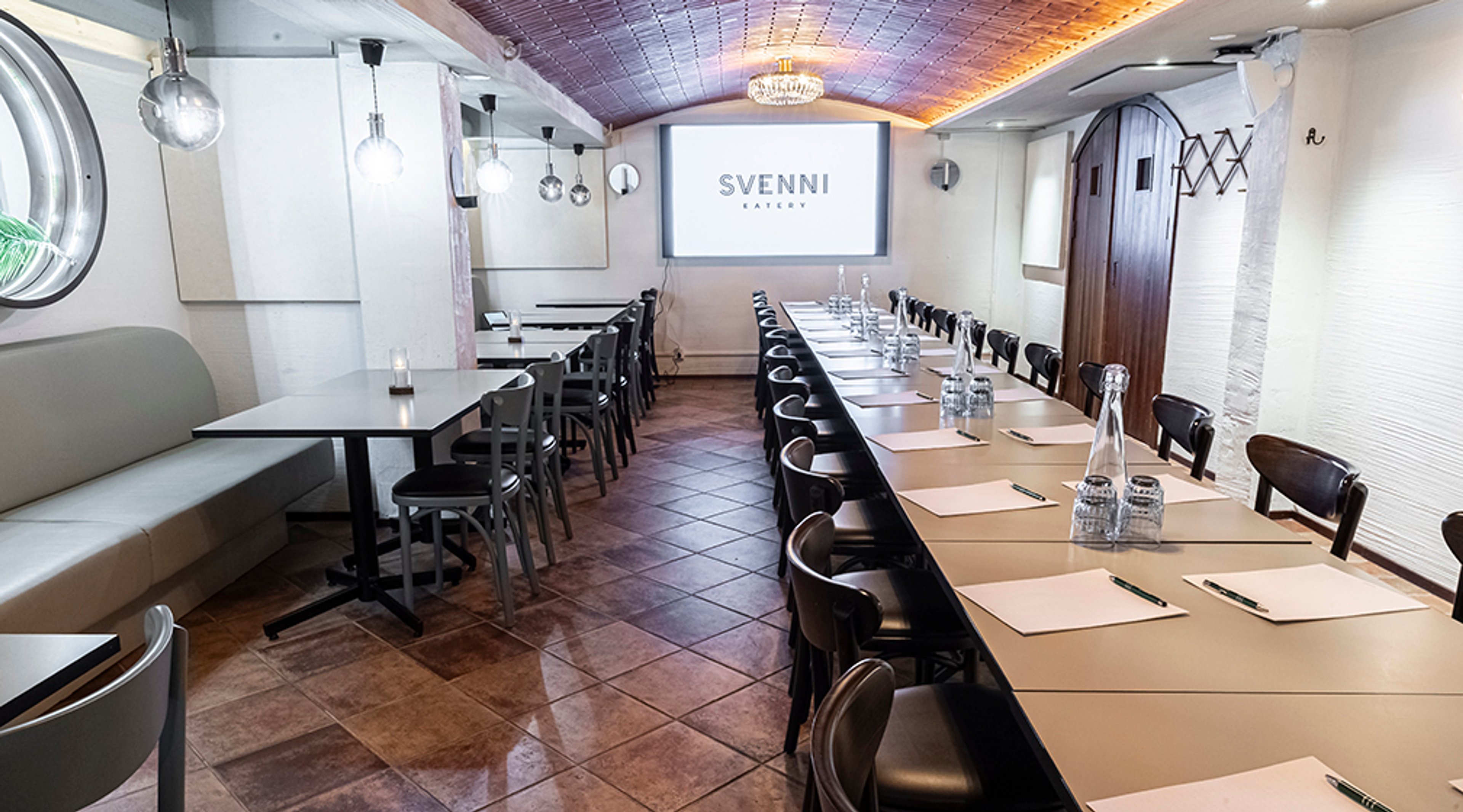 Kokoustila, jossa pitkä pöytä tuoleineen, seinäpenkki ja pienempiä pöytiä. Tilassa on tasainen lattia ja valaistus kattokruunusta.