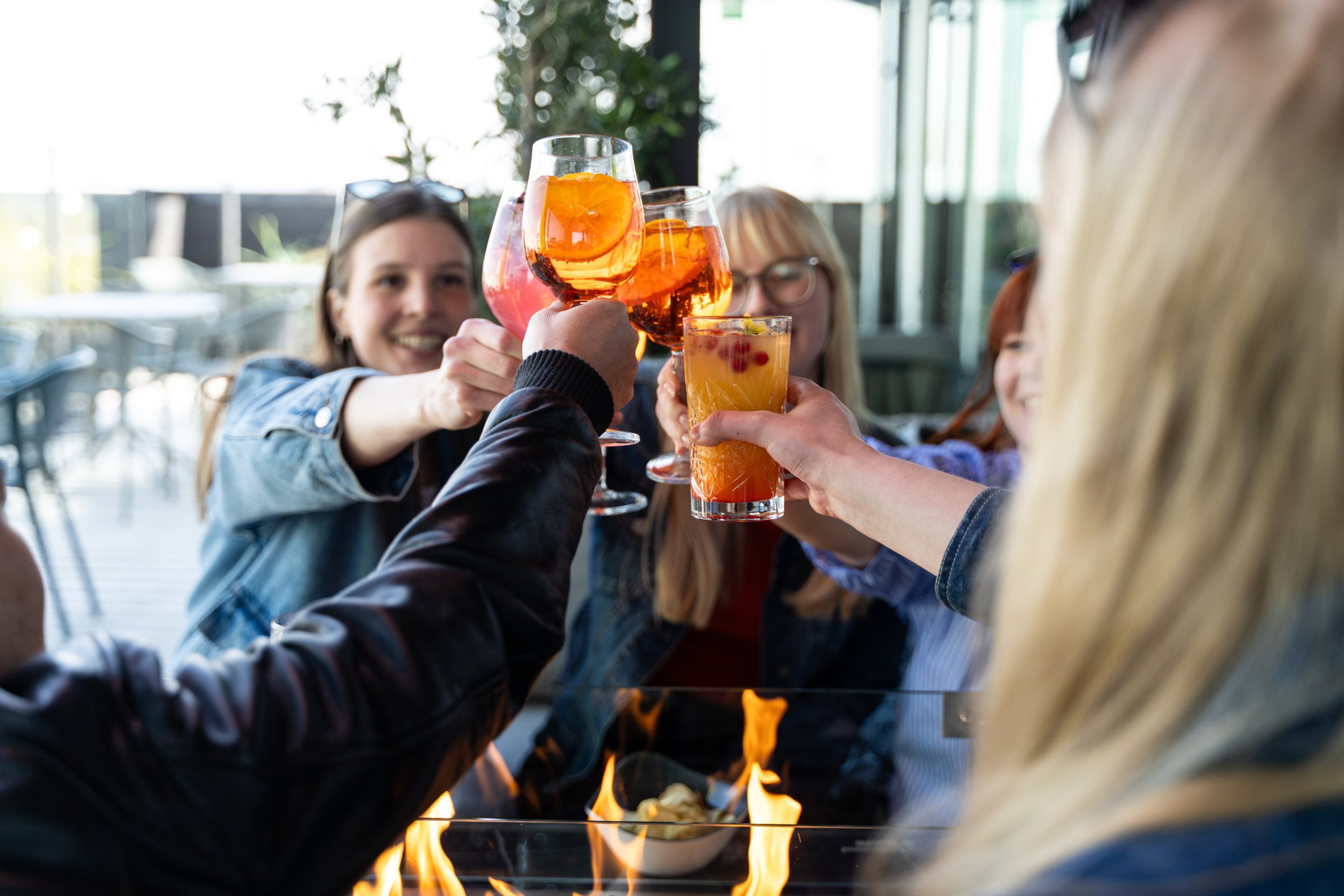 Ryhmä henkilöitä kohottaa värikkäitä juomia maljaksi pöydän ääressä, jossa on pieni tuli tai liekki etualalla.