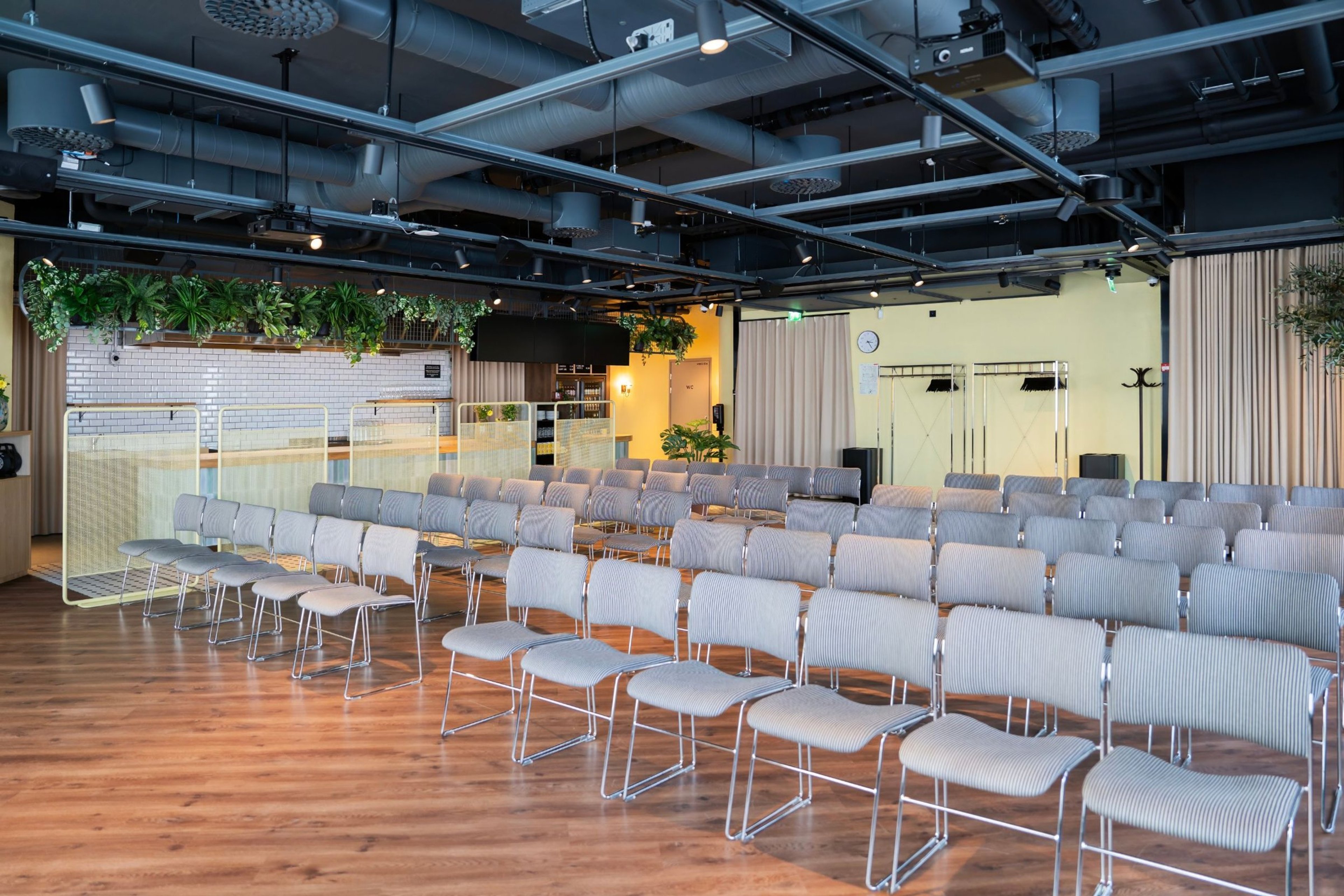Moderni auditorio riveittäin aseteltuja harmaita tuoleja, puulattia, valkoinen tiiliseinä ja viherkasvikoristelu. Avara tila, jossa esteetön kulku.