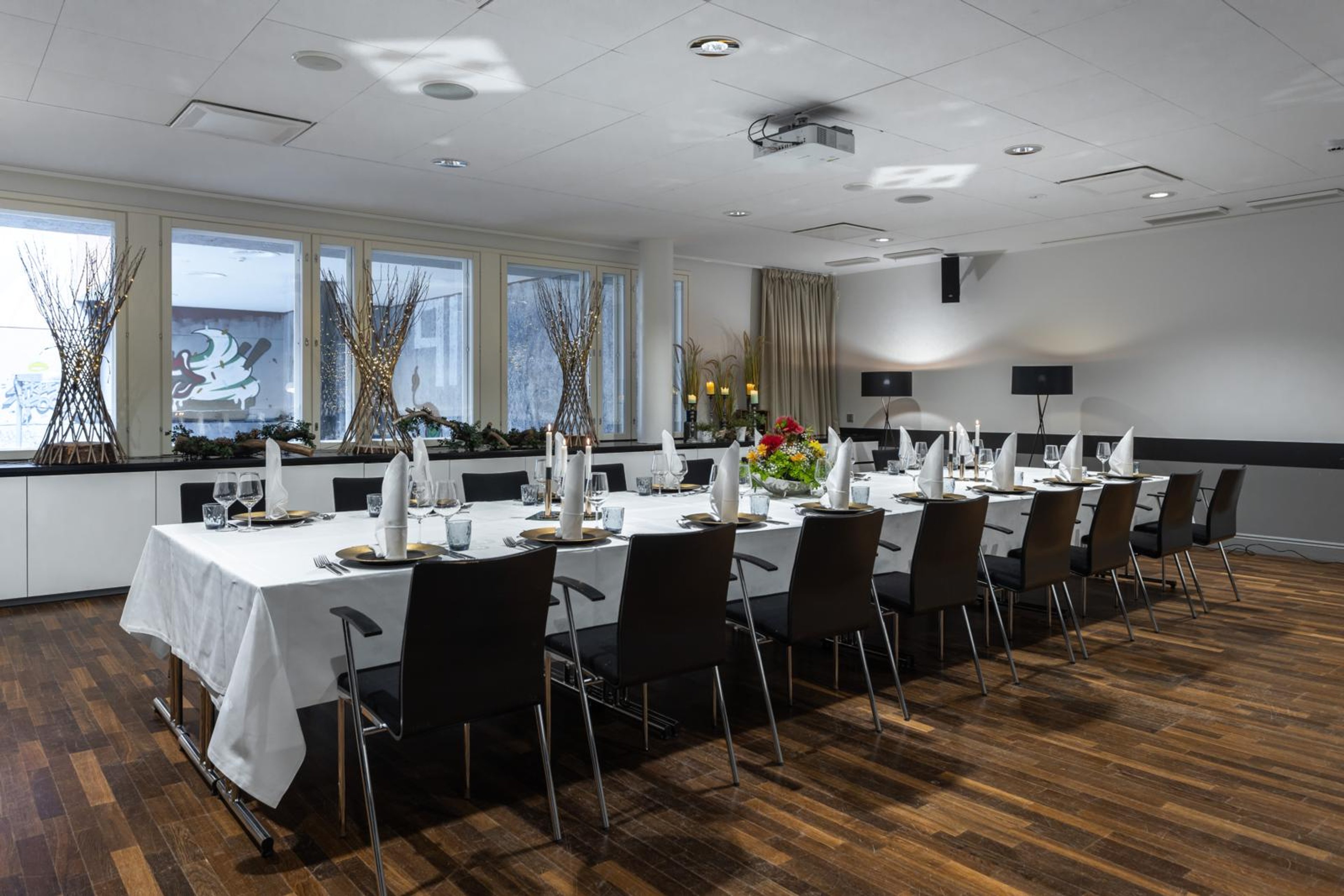 Kokoustila jossa pitkä pöytä valkoisen pöytäliinan alla. Tummat tuolit, puulattia ja ikkunoilla koristerisuja. Katossa projektori.