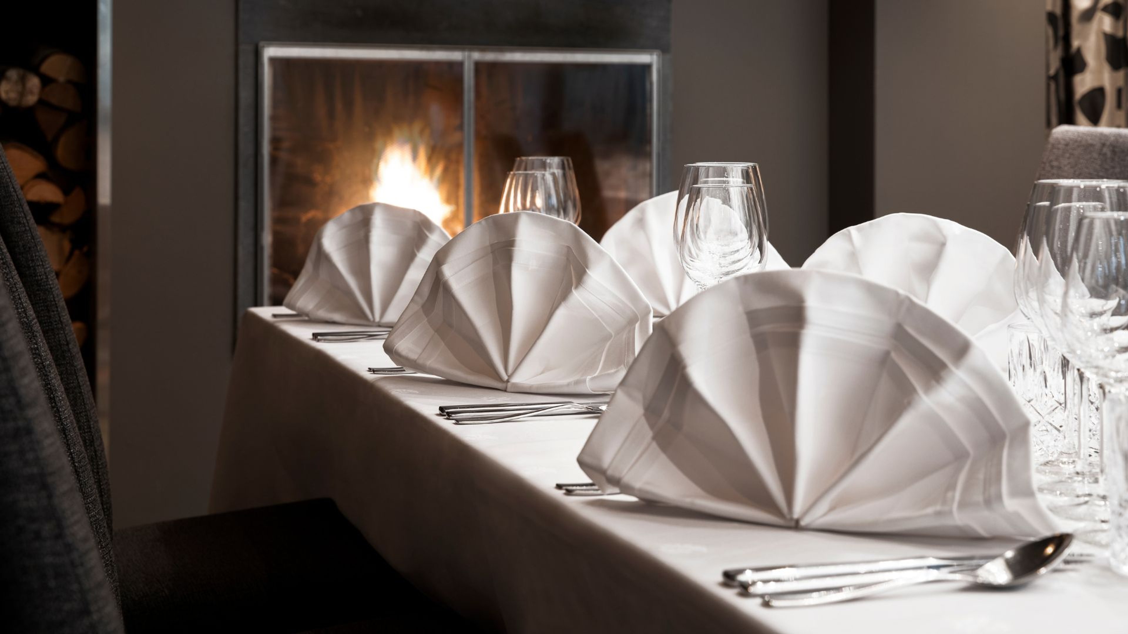 Elegantisti katettu pöytä taitelluin lautasliinoina ja viinilaseilla, taustalla loimuaa takkatuli luoden tunnelmallisen ilmapiirin.