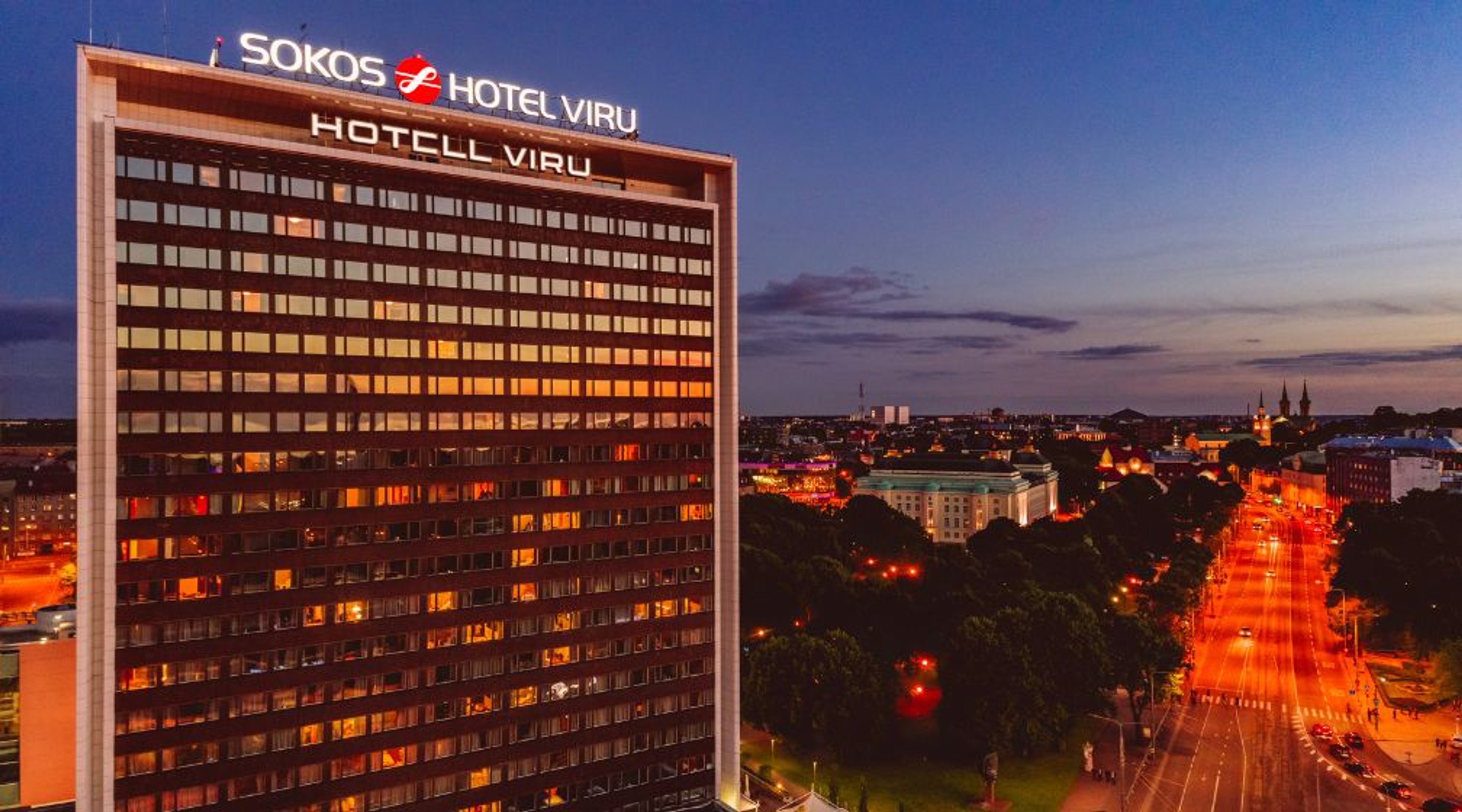 Valaistuja ikkunoita omaava korkea hotellirakennus iltahämärässä, ympäröivä kaupunkinäkymä.