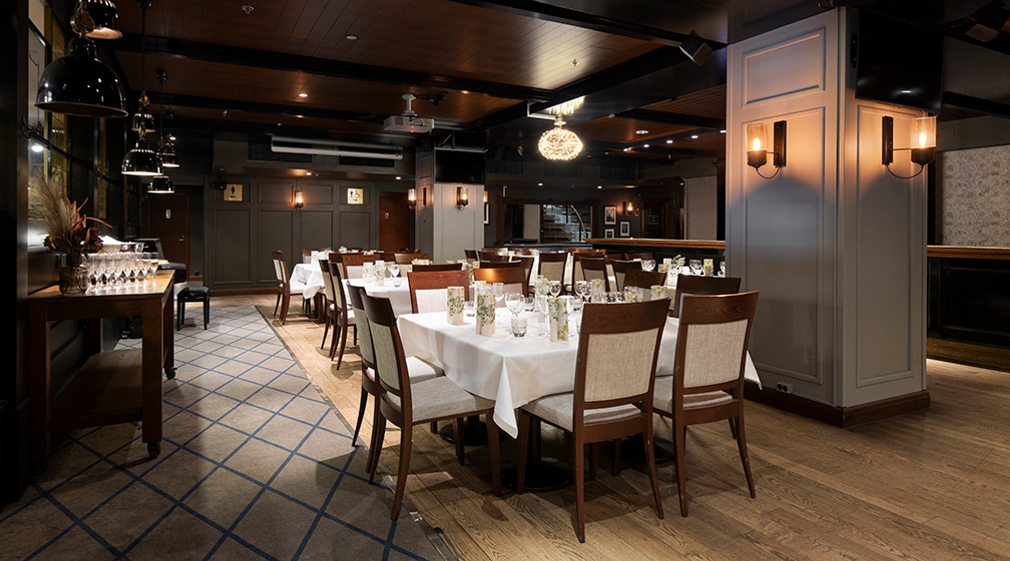 Tyylikkäästi sisustettu ravintolasali puulattialla, pöydissä valkoiset pöytäliinat ja puiset tuolit. Tila on valaistu tunnelmallisesti.