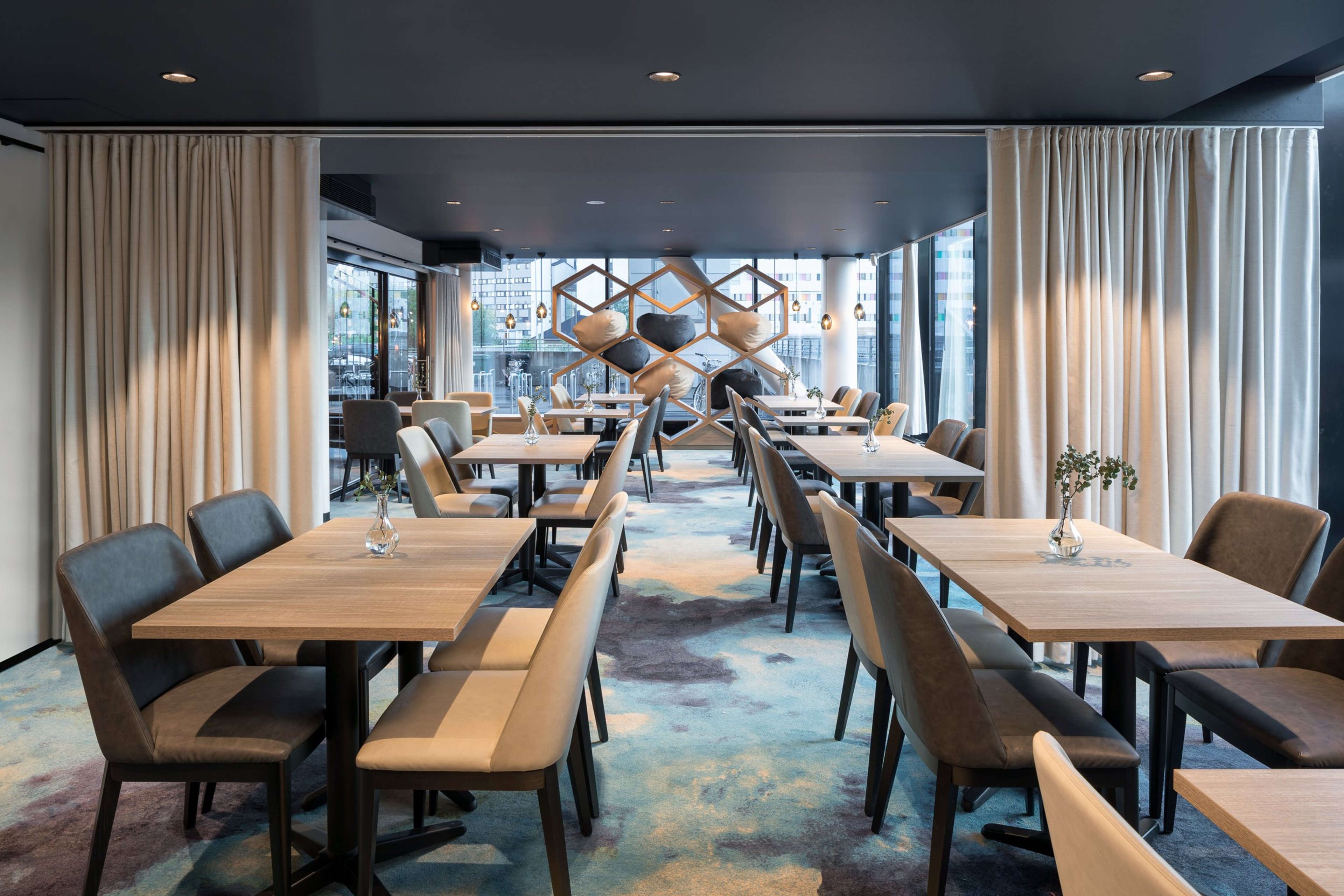 Moderni ravintola, jossa puiset pöydät, pehmeät tuolit, iso lattiasta kattoon ulottuva ikkuna ja sisustustaulut. Tasainen lattia ja runsas valaistus.