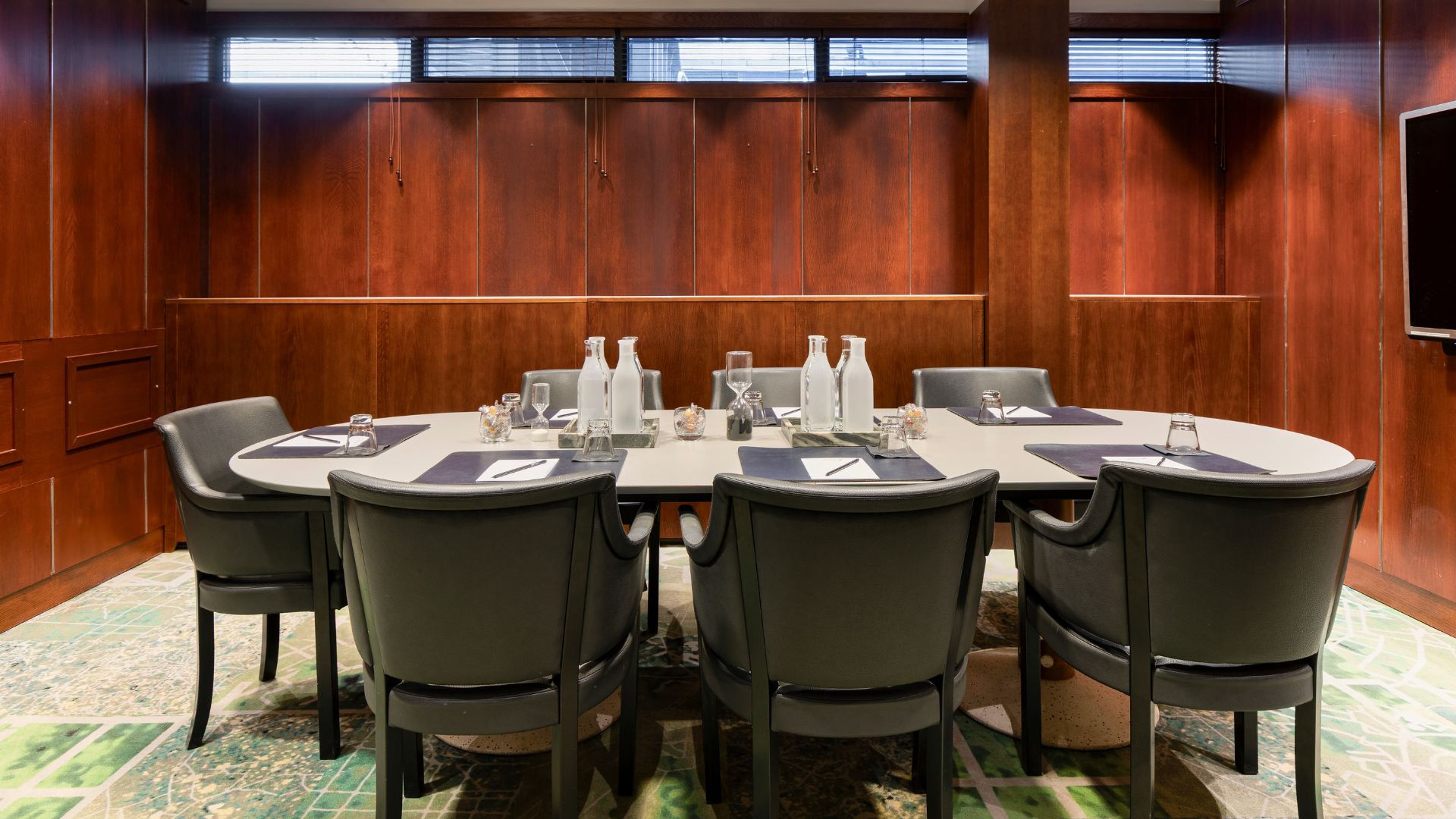 Kokoushuone puupaneeliseinillä ja valkoinen pöytä mustilla tuoleilla. Pöydällä on vesipulloja ja laseja patterneilla.