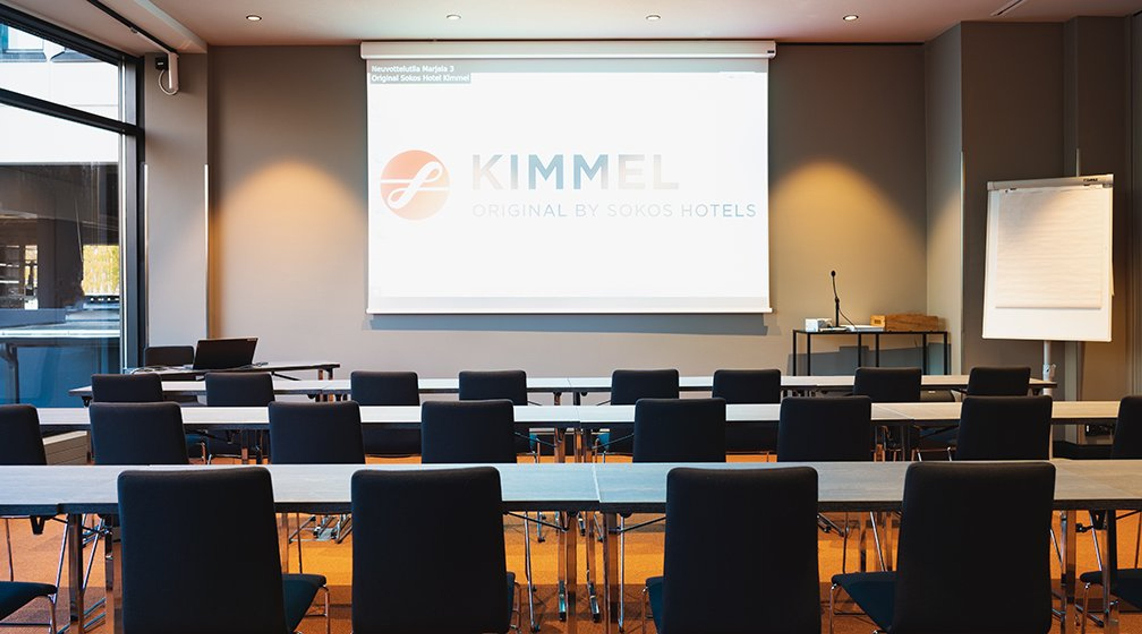 Kokoustila vaaleilla pöydillä ja tummilla tuoleilla rivissä. Edessä valkokangas, suuret ikkunat ja tasainen puulattia.