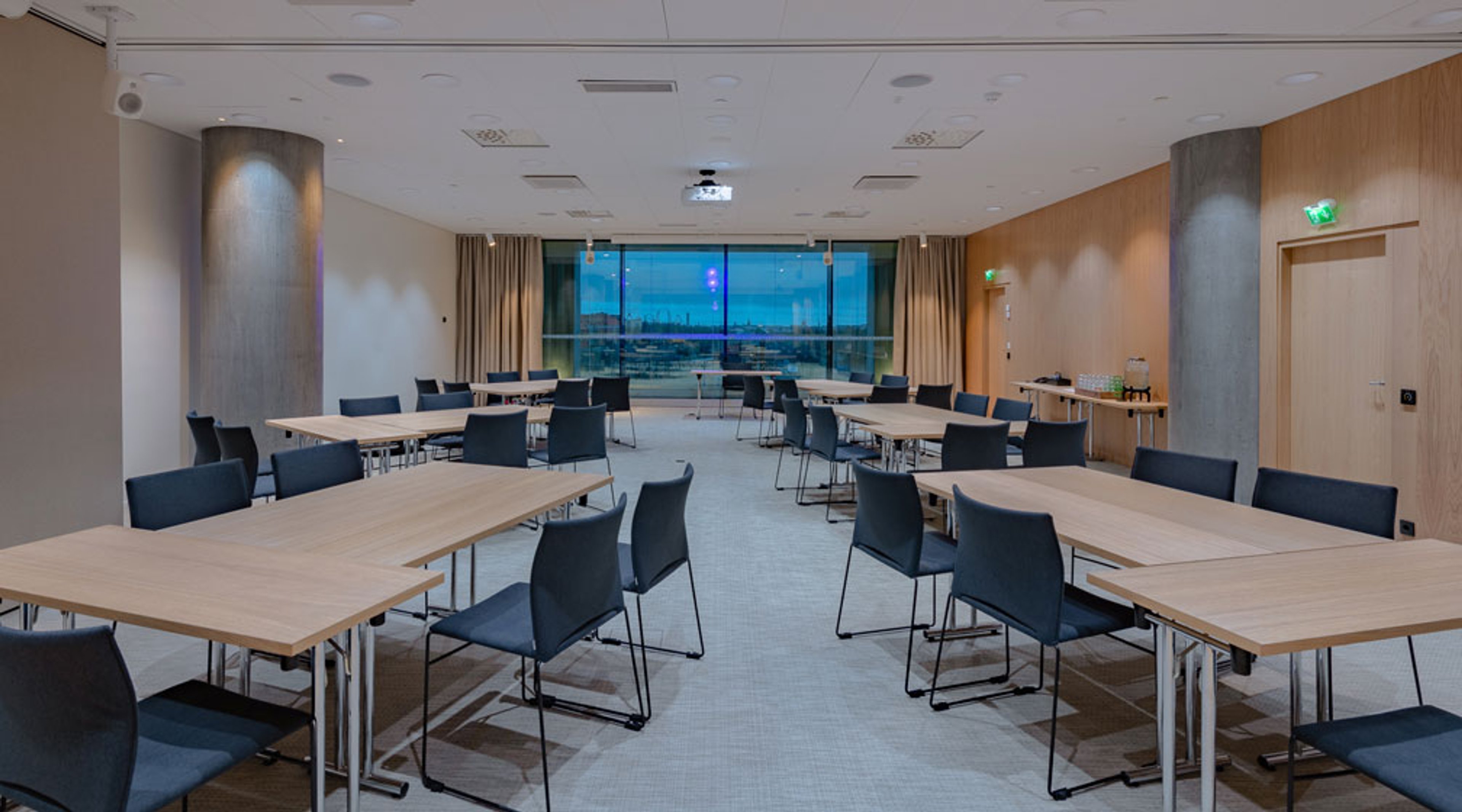 Kokoushuone, jossa puiset pöydät mustat tuolit U-muodostelmassa. Takana lasinen seinä, josta näkyy maisema. Tasainen lattiapinta ja leveät kulkutilat.