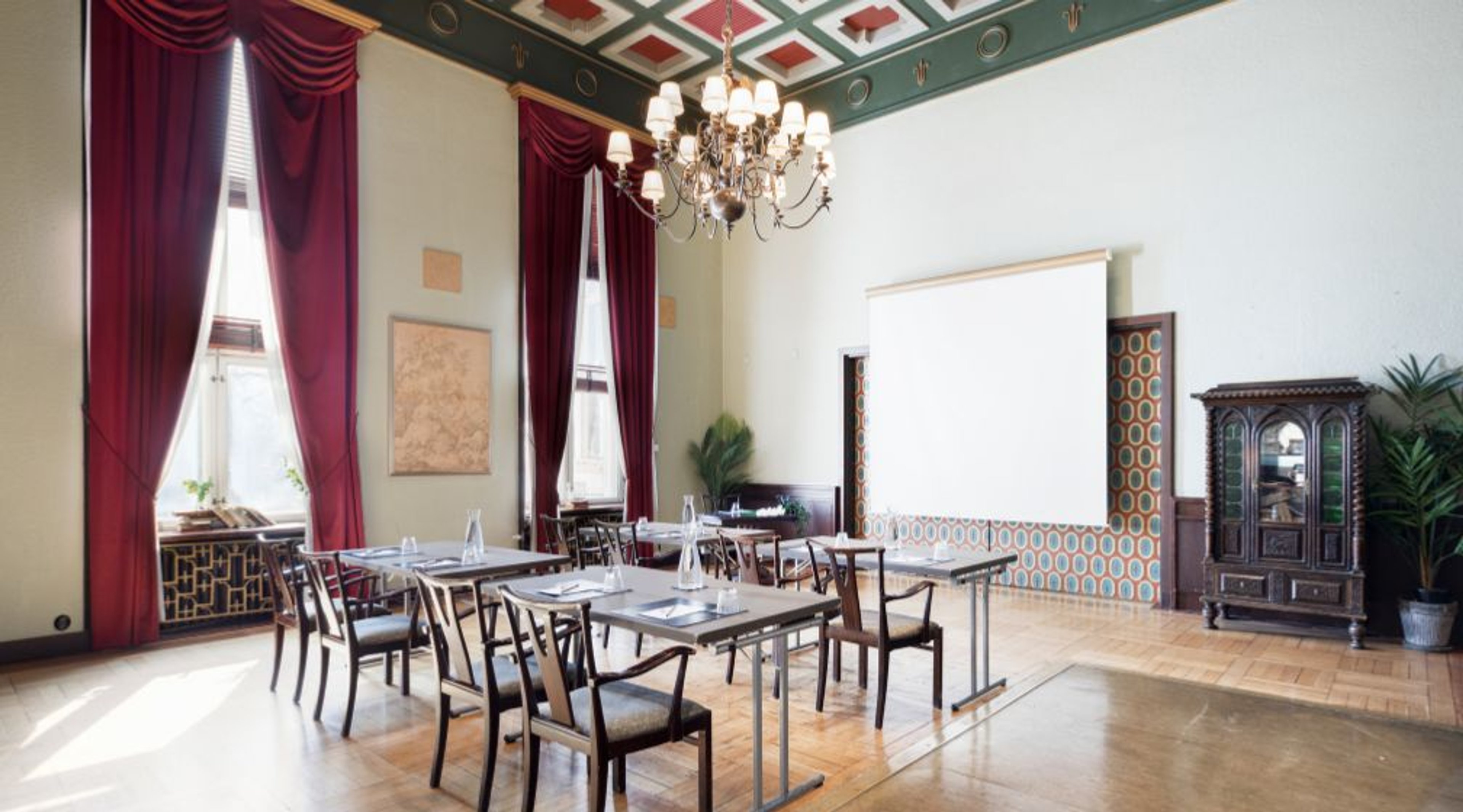 Ballroom with high decorated ceiling, crystal chandelier, tall windows with burgundy curtains and wooden floor. The space features tables and chairs arranged in conference style.