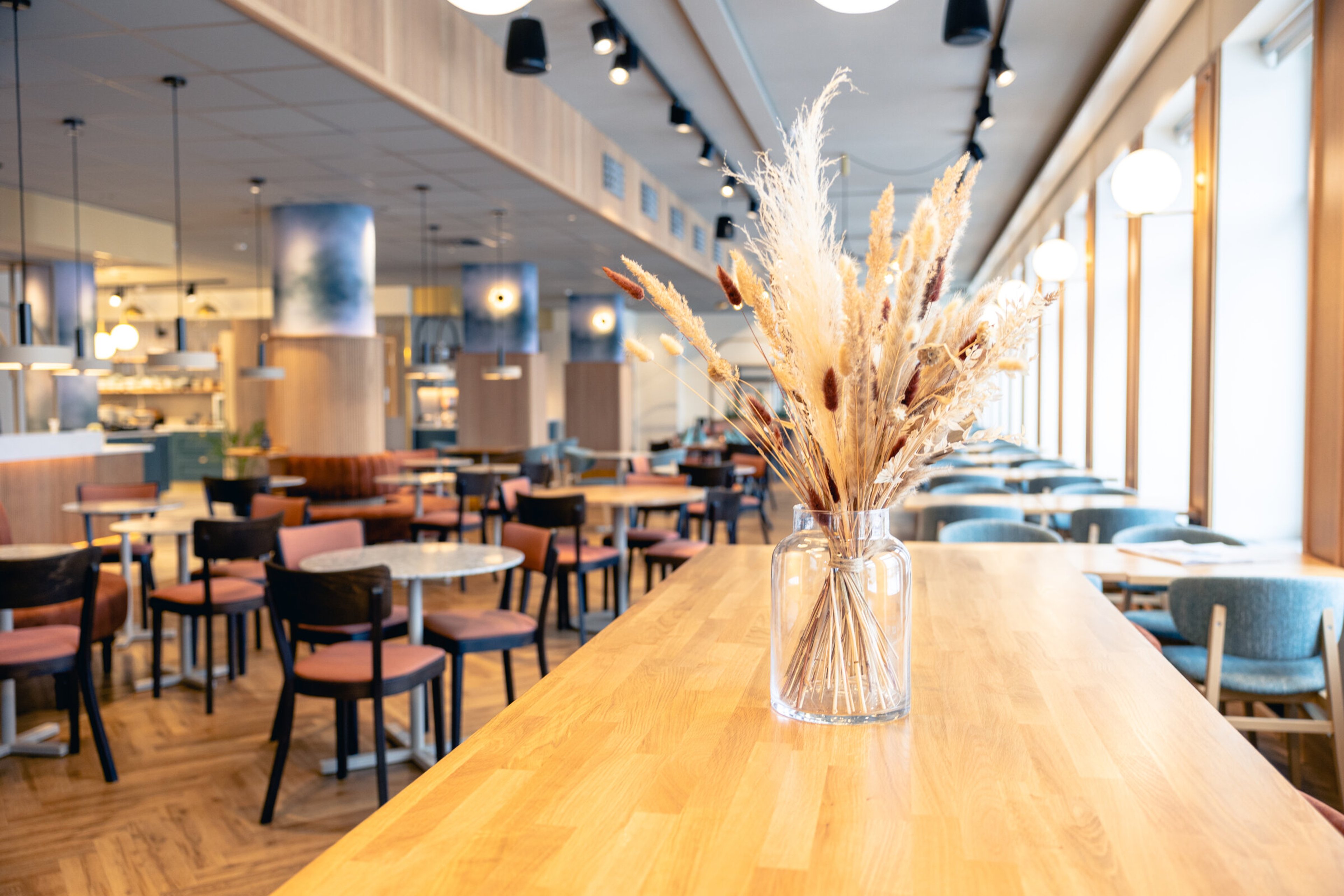 Modern restaurang med ljust trägolv, olika sittplatser och gott om utrymme att röra sig. Stora fönster ger rikligt med naturligt ljus.