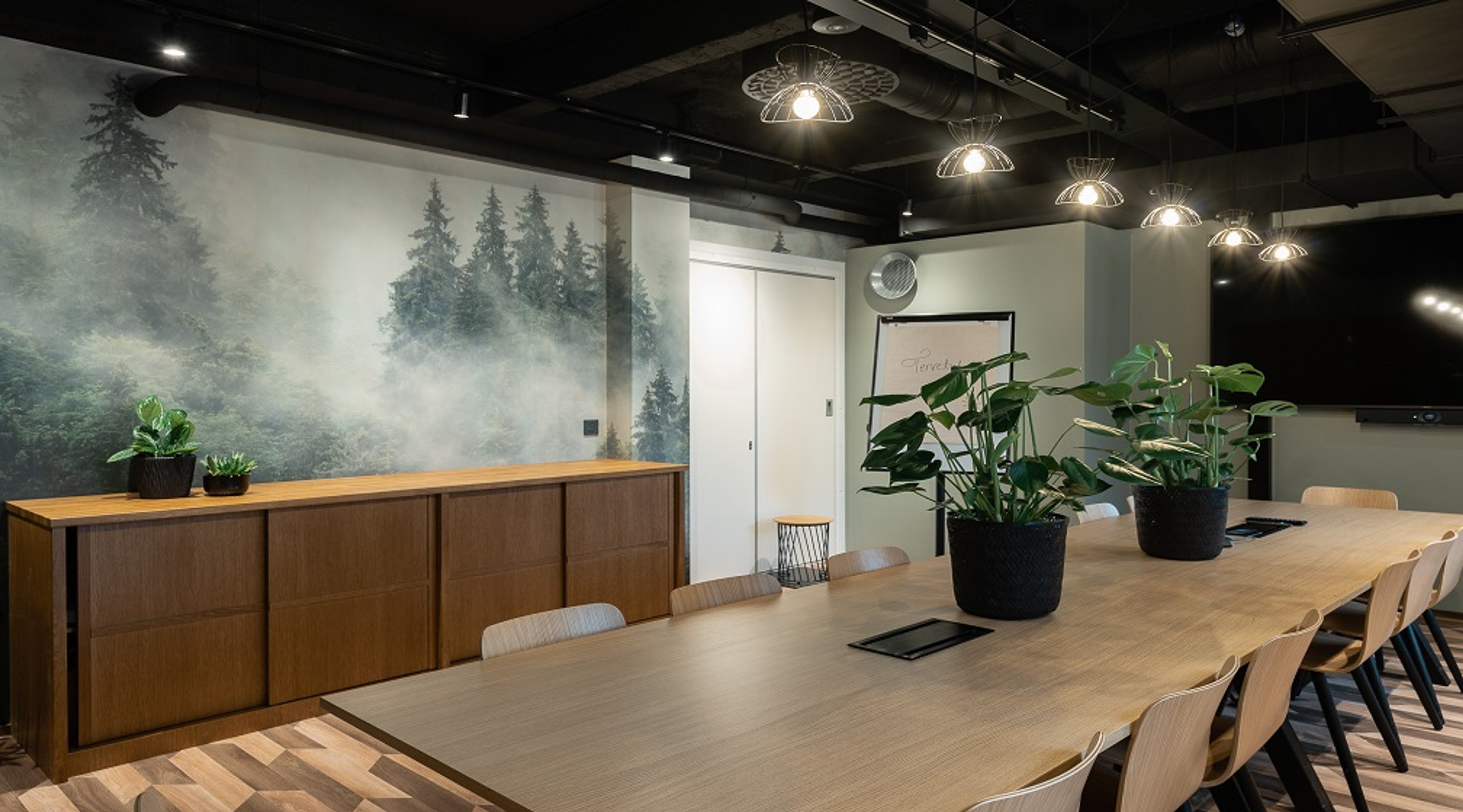 Moderni neuvotteluhuone, jossa puinen pöytä, huonekasveja ja suuri metsäaiheinen seinämaalaus.
