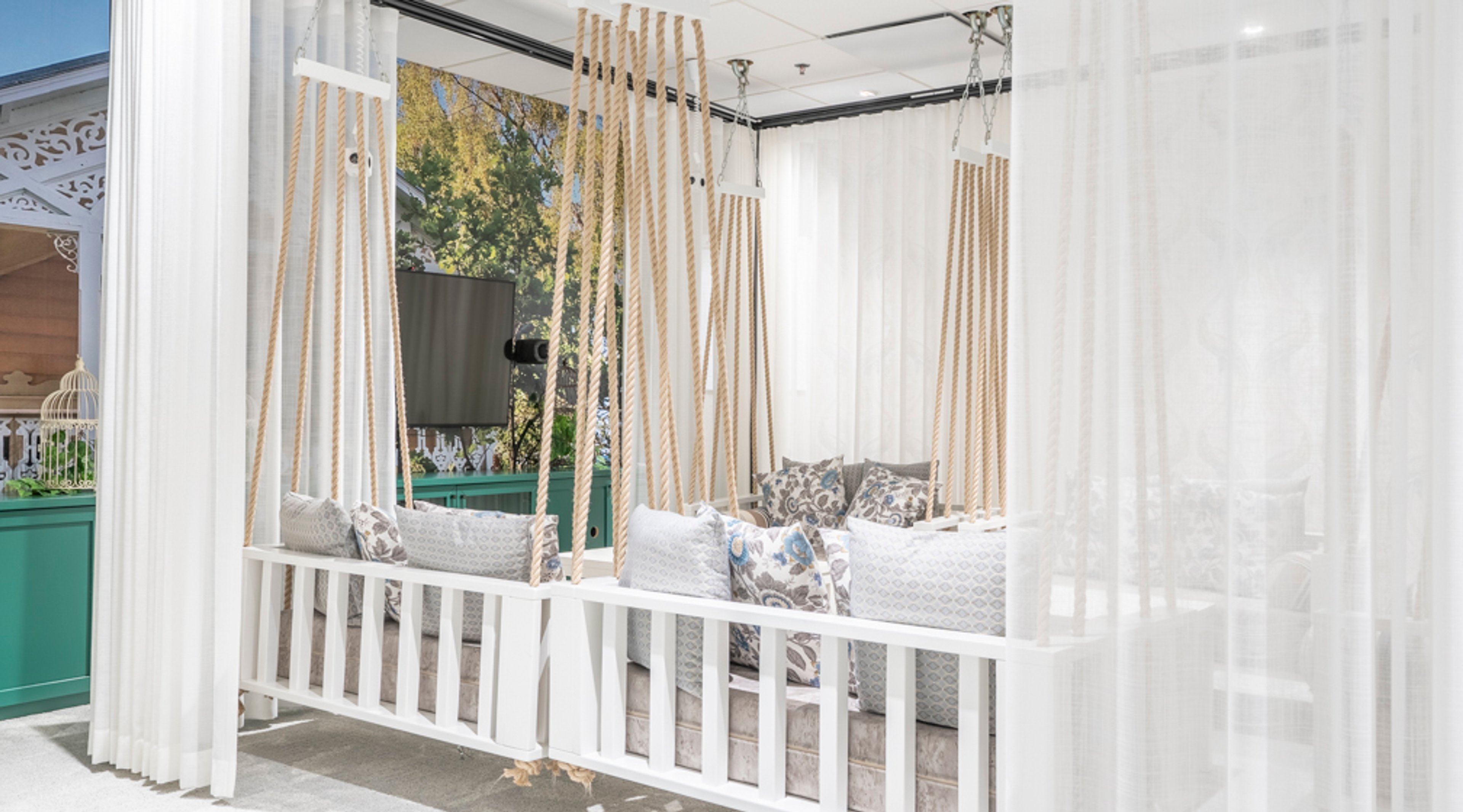 Sisätilojen keinu-istuinryhmä valkoisilla verhoilla, köysikiinnityksellä ja runsailla tyynyillä. Taustalla näkyy ikkuna ja televisio.