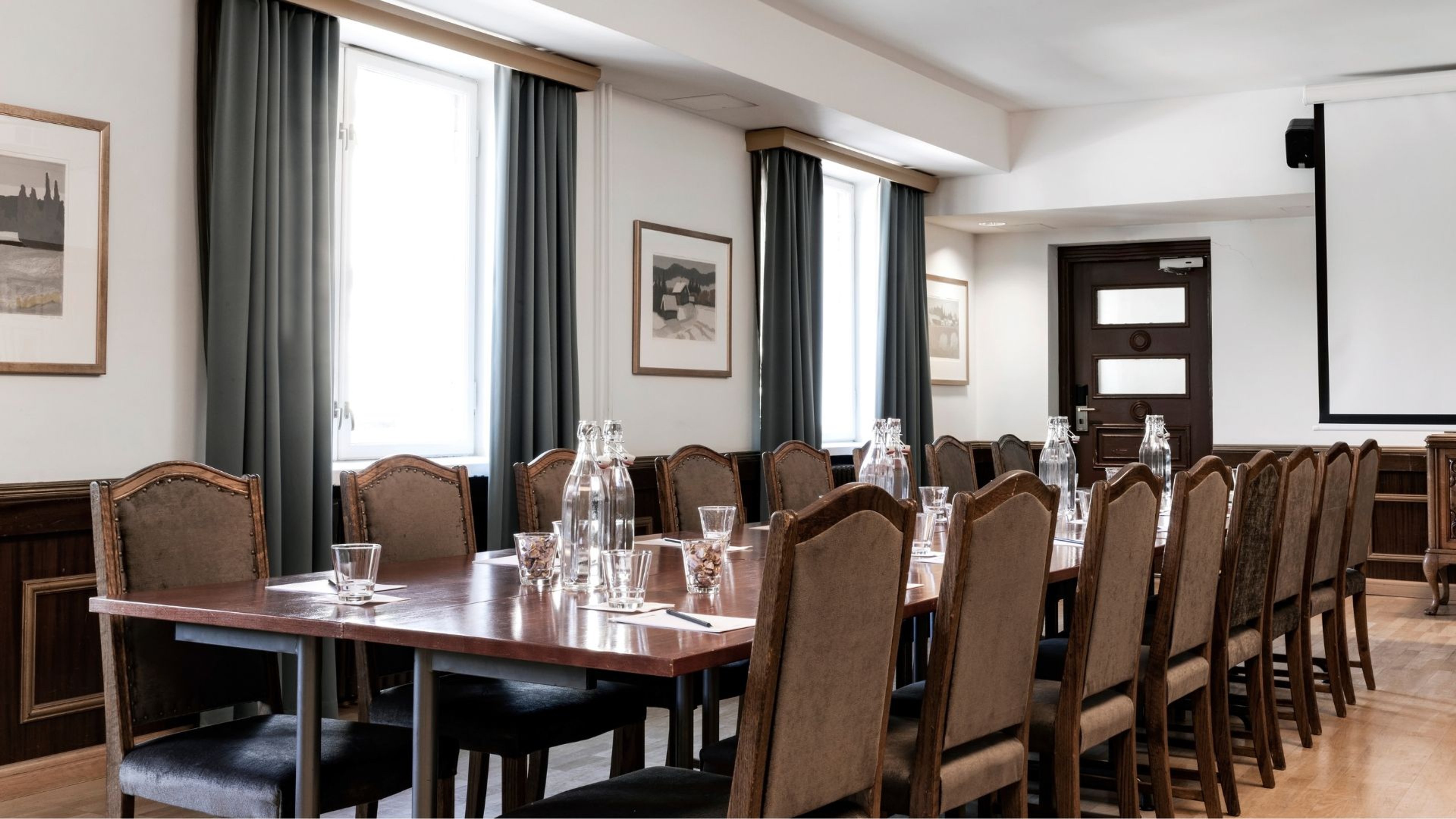 Kokoushuone, jossa suuri pöytä, upholsteroidut tuolit ja valkoinen projektoriruutu. Huoneessa kaksi suurta ikkunaa vihreillä verhoilla ja puulattia.