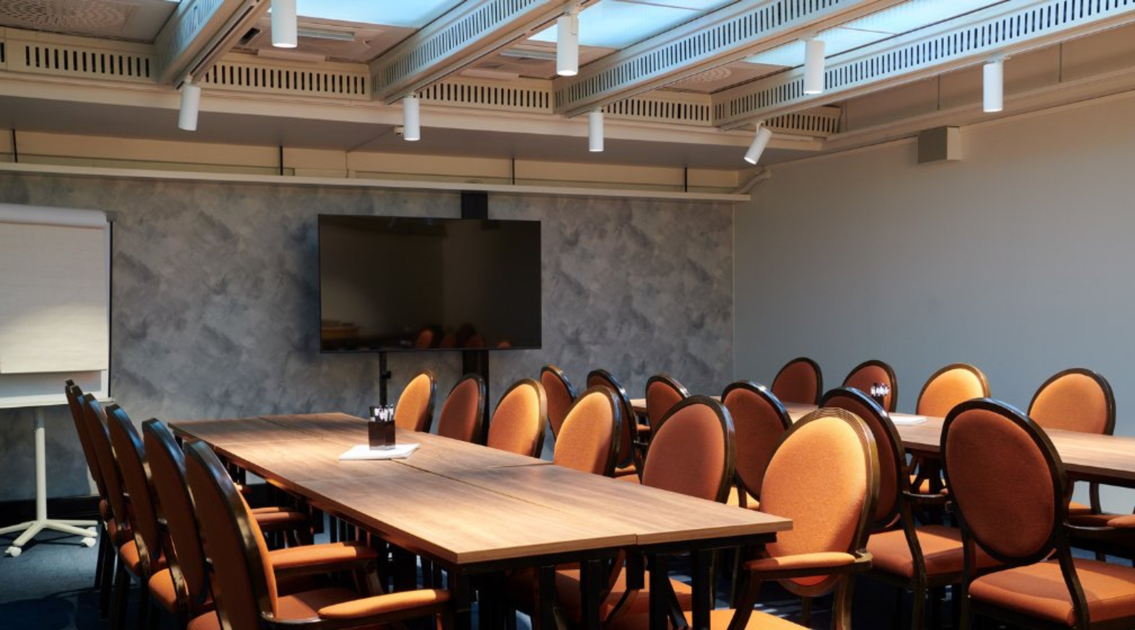 Kokoustila, jossa puupöytiä ja oranssit tuolit. Seinällä suuri näyttö ja taustalla vaalea marmoriseinä sekä valkoinen katto.