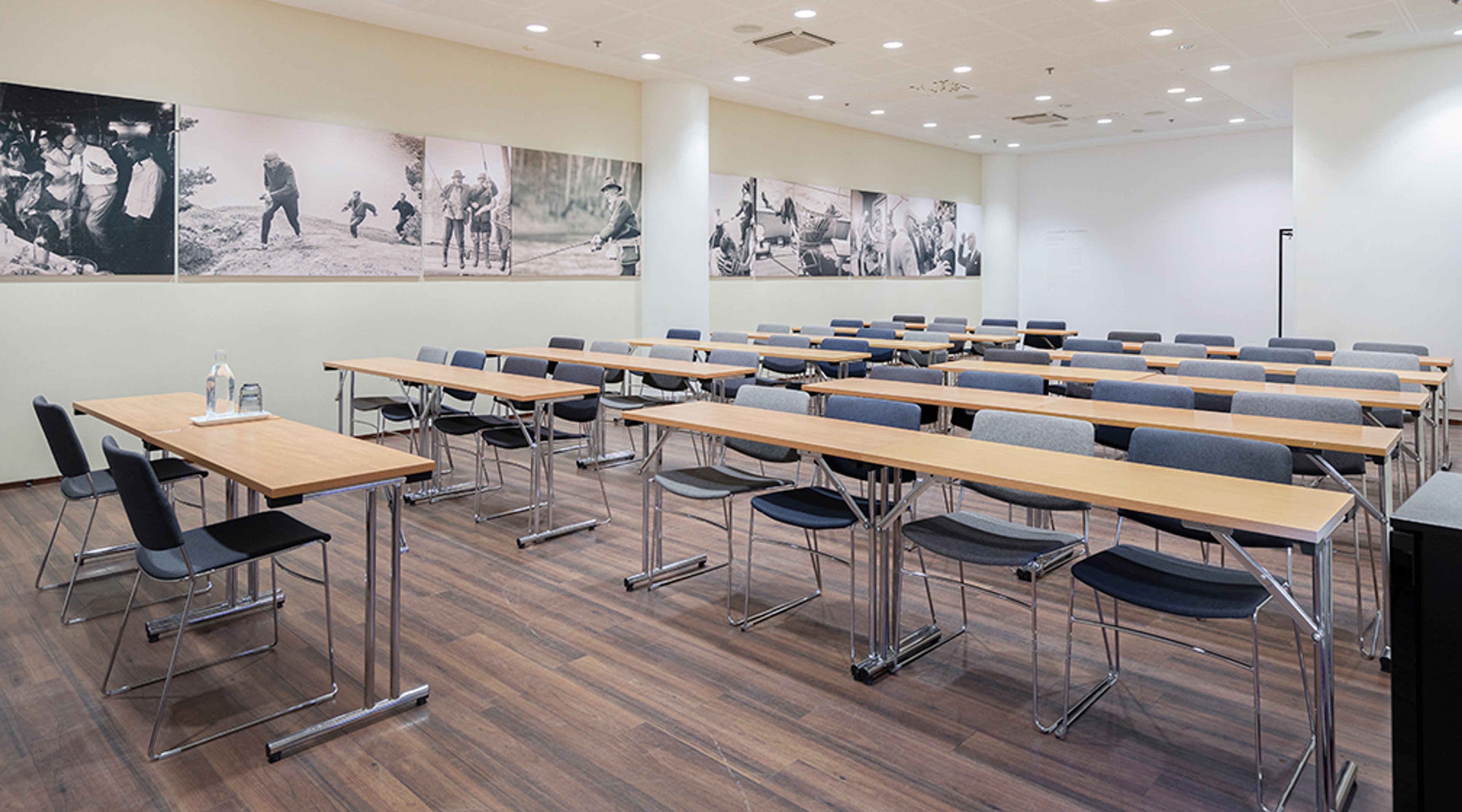 Kokoustila puisilla pöydillä ja siniharmailla tuoleilla. Seinällä historiallisia valokuvia. Tasainen puulattia, esteetön kulku.