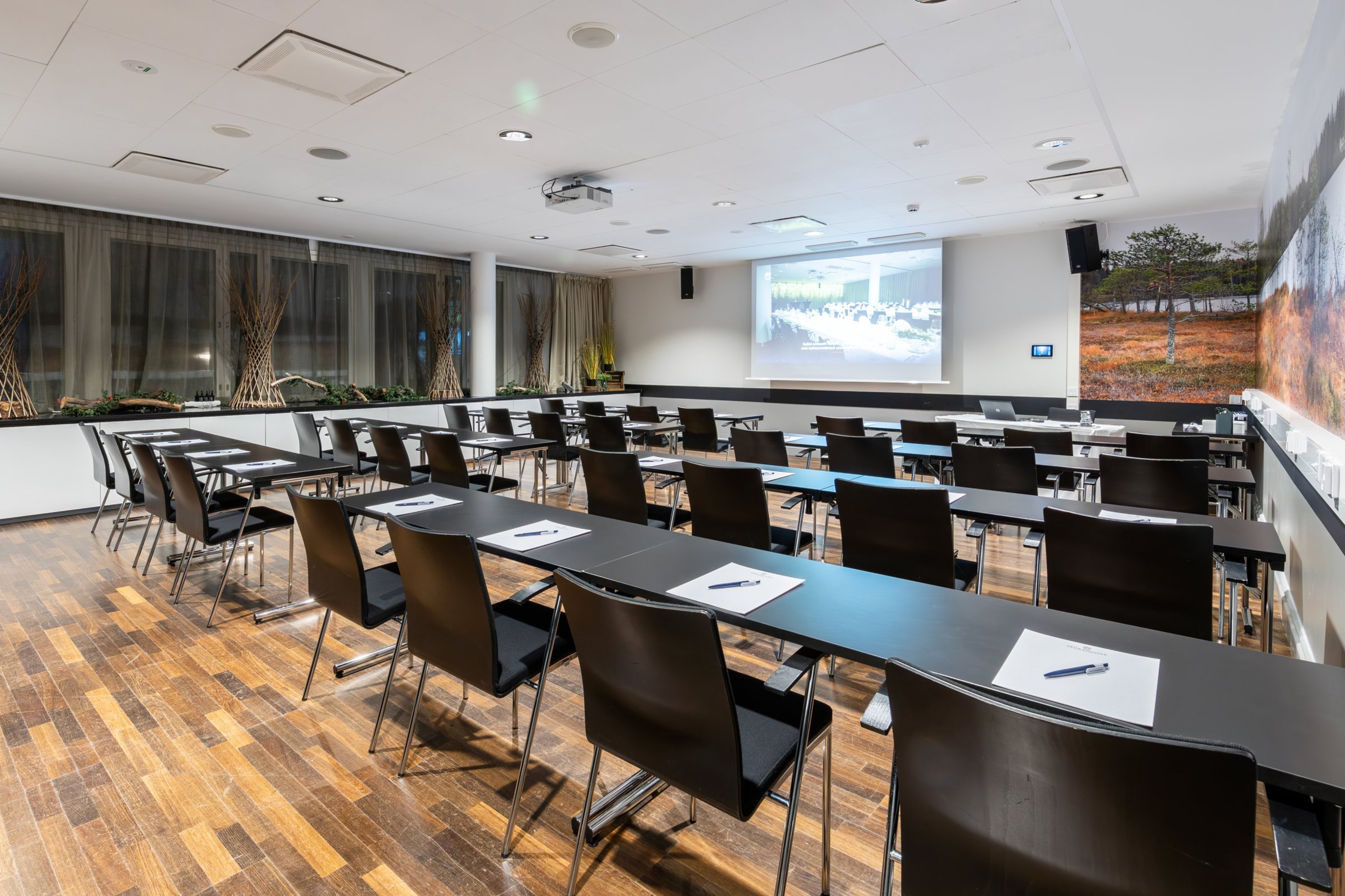 Valoisa kokoustila pitkillä pöydillä ja mustilla tuoleilla, puulattia, seinällä valkokangas ja luontomaalaus.