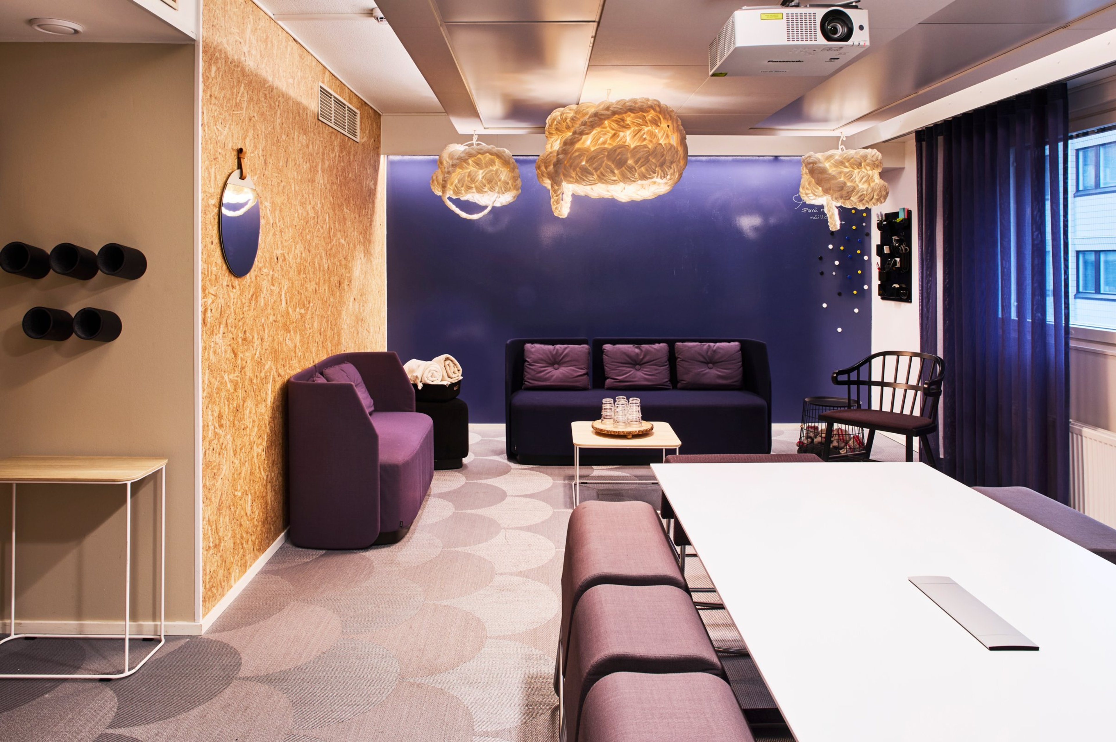 Moderni neuvotteluhuone, jossa violetti sohva, valkoinen pöytä, projektori katossa ja koristeelliset valaisimet. Lattia on pehmeä kuvioitu matto.