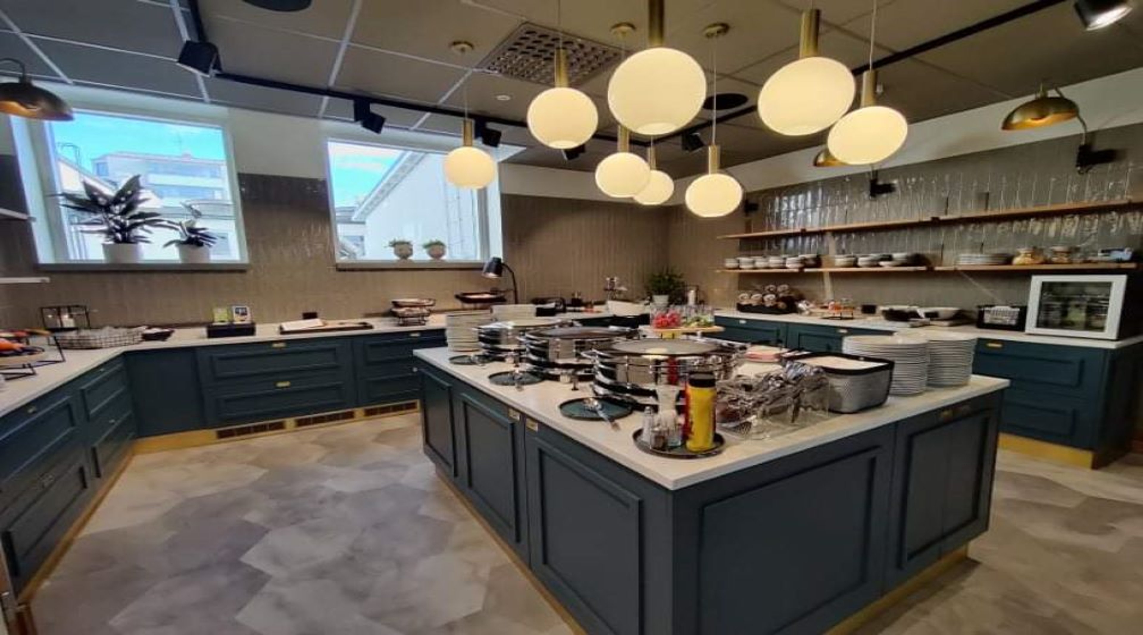 Moderni buffetpöytä sinisillä kaapeilla, valkoisilla pöytätasoilla ja pyöreillä riippuvalaisimilla. Tilassa suuret ikkunat ja avarat käytävät.