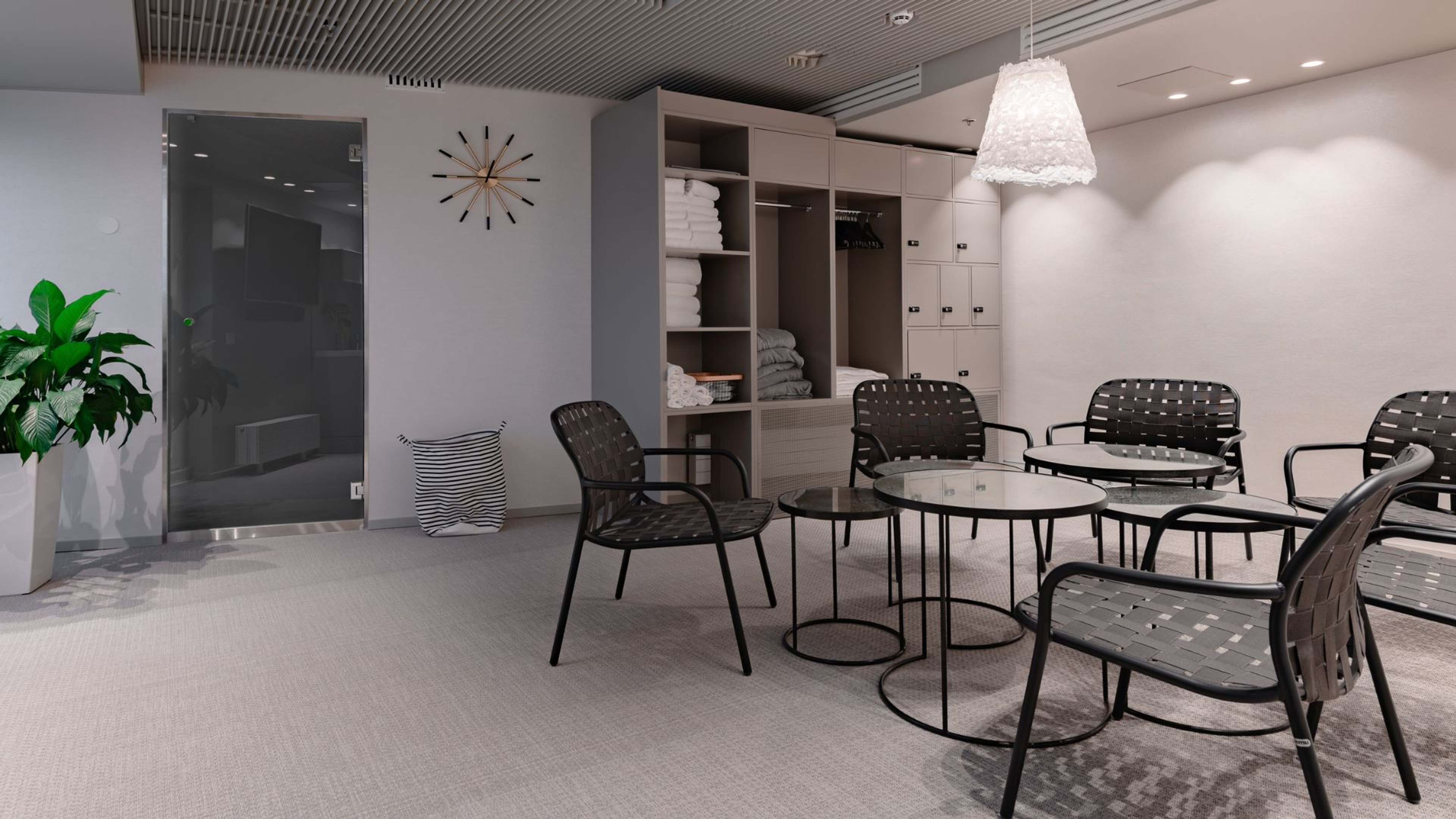 Moderni saunatilan odotushuone, jossa on mustia tuoleja, pyöreitä pöytiä, pyyhehylly, lasinen saunan ovi ja kasveja. Tasainen lattiapinta ja hyvä valaistus.