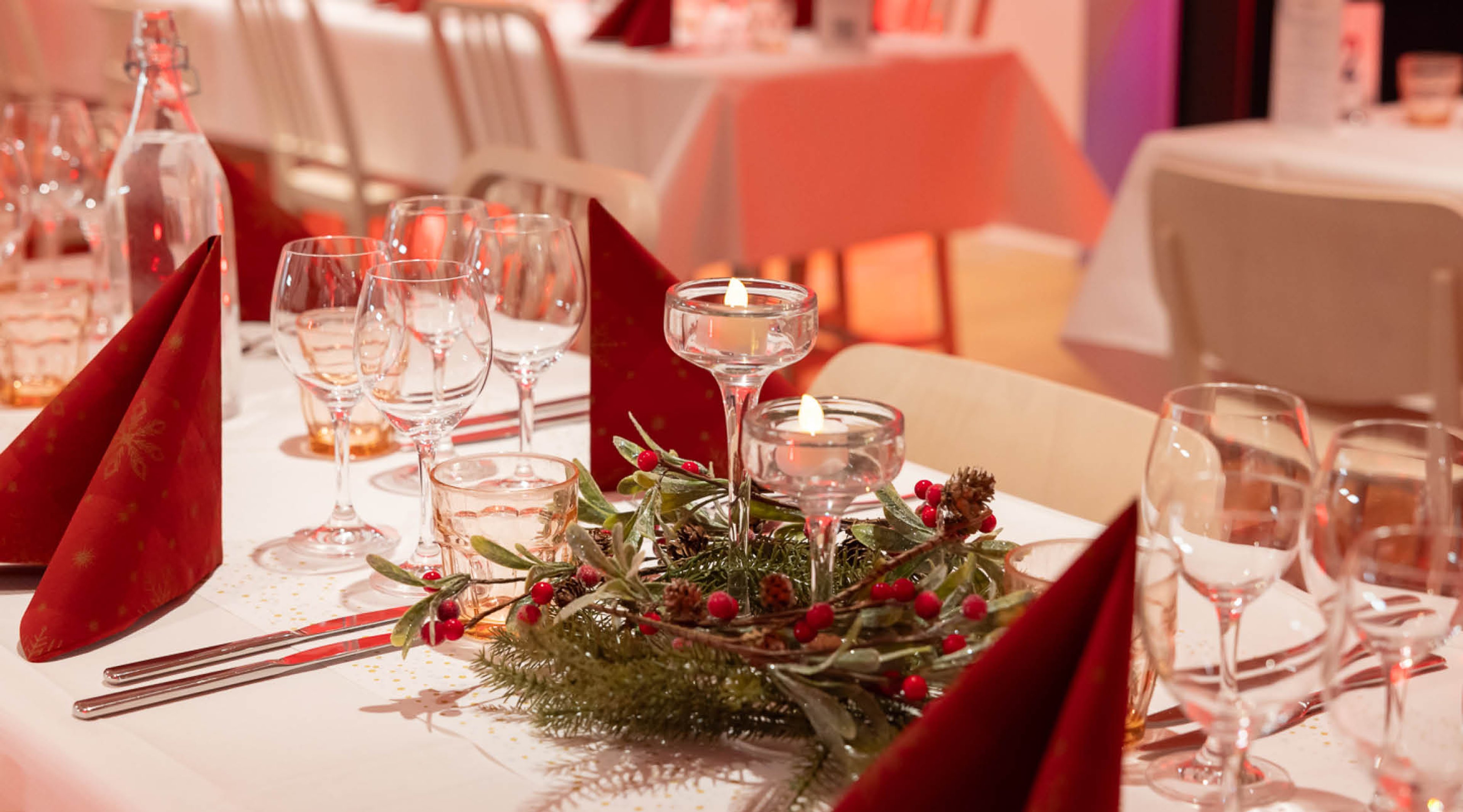 Juhlallisesti katettu pöytä, jossa punaisia lautasliinoja, kynttiläkoristeita ja joulunpunaisia marjoja havukoristeessa.