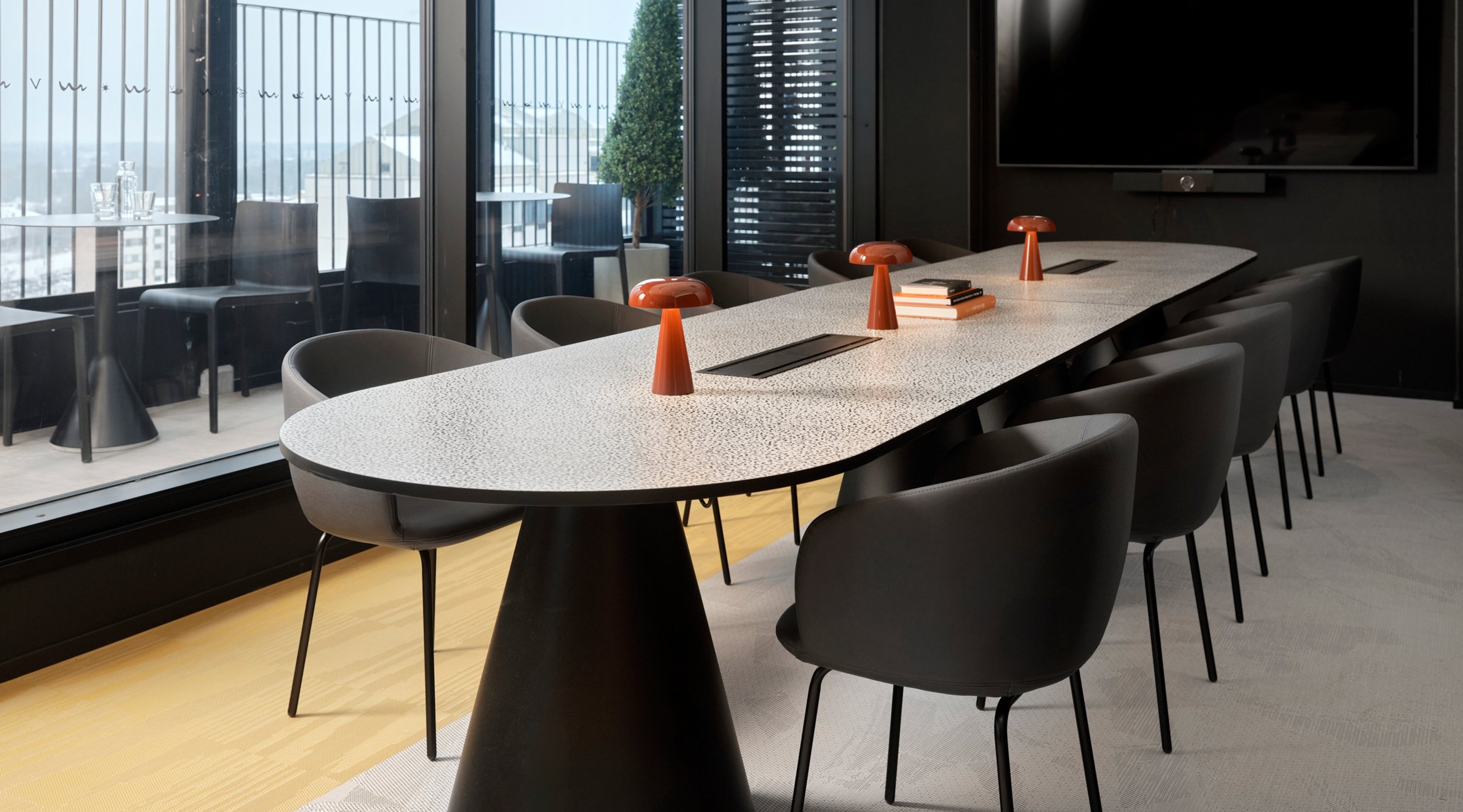 Moderni neuvotteluhuone, jossa valkoinen pöytä ja mustat tuolit. Oranssit pöytävalaisimet ja isot ikkunat tuovat luonnonvaloa. Puulattia ja TV-näyttö seinällä.