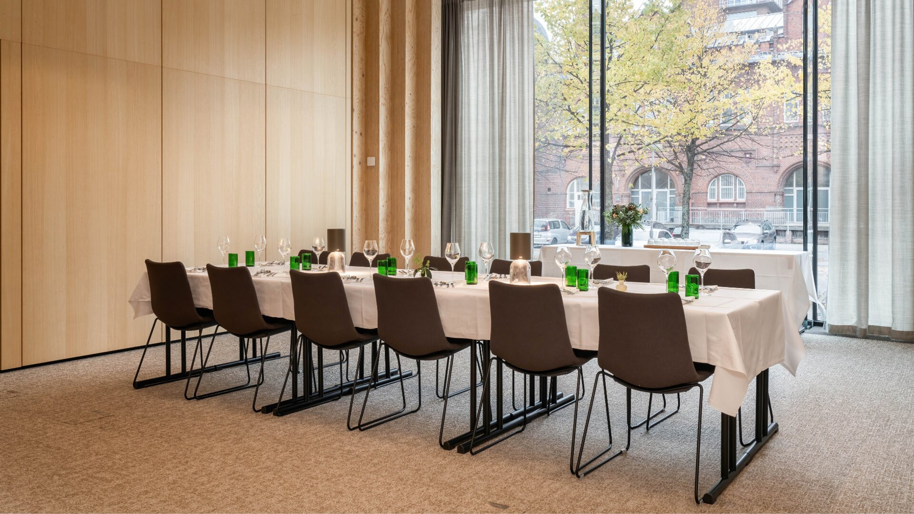 Konferensrum med långt bord täckt av vit duk och bruna stolar. Stora fönster visar träd och en tegelbyggnad utanför. Jämn heltäckningsmatta.