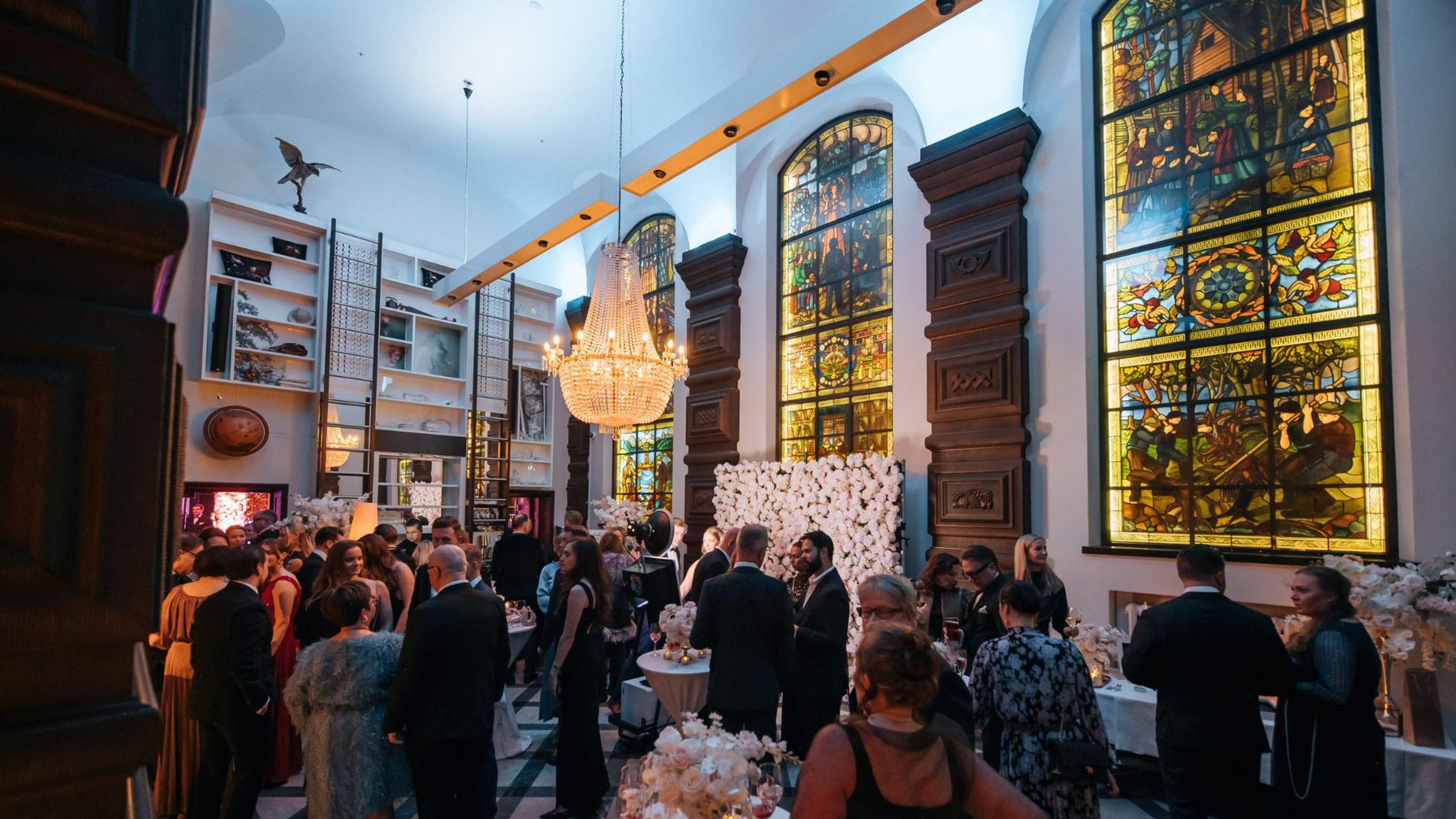 Juhlasali koristeellisilla lasimaalauksilla, kristallikruunulla ja valkoisilla kukkakoristeilla, jossa elegantisti pukeutuneita vieraita.