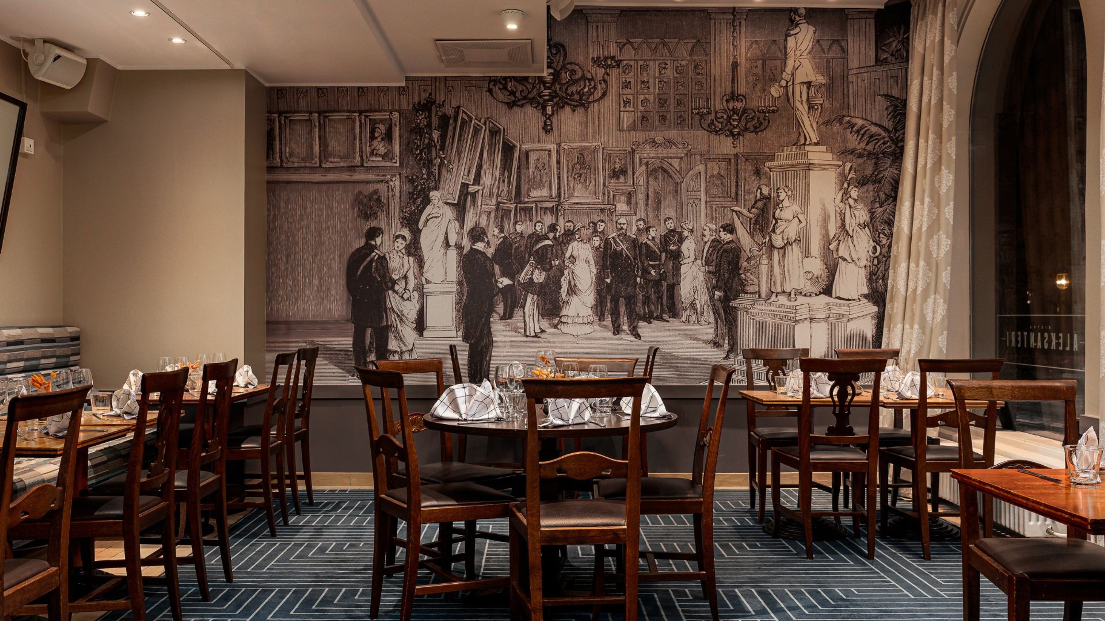 Ravintolan sisätila, jossa puisia pöytiä ja tuoleja. Taustalla suurikokoinen historiallinen seinämaalaus. Kuvioitu lattia ja tasainen valaistus.