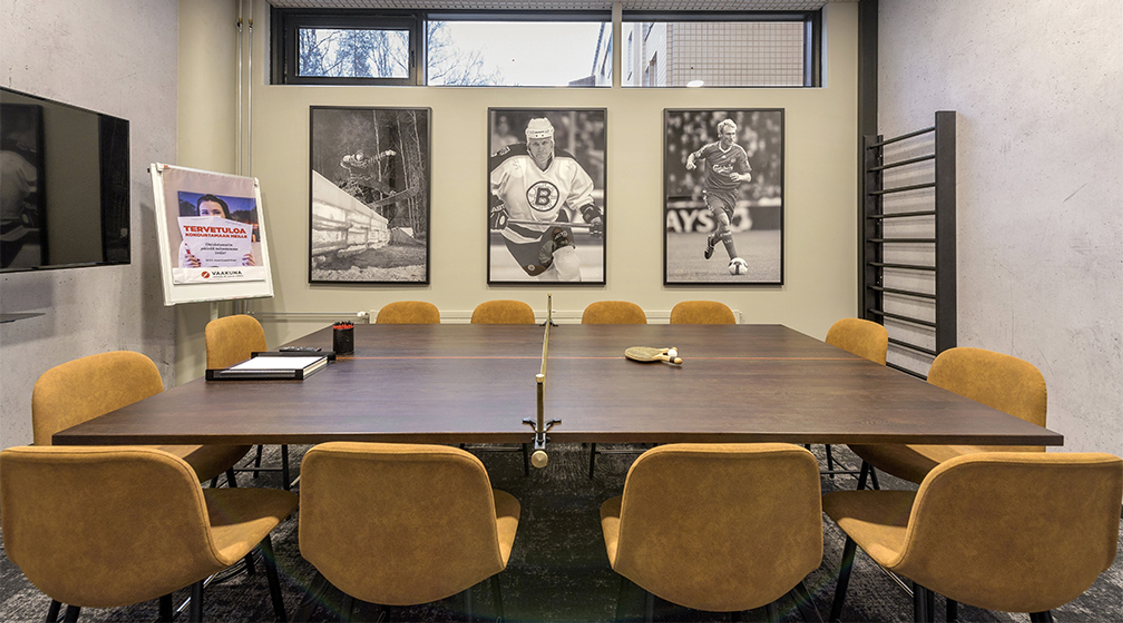 Neuvotteluhuone, jossa iso pöytä pingispeliin muunnettuna. Seinällä urheiluaiheisia valokuvia ja keltaiset tuolit pöydän ympärillä.