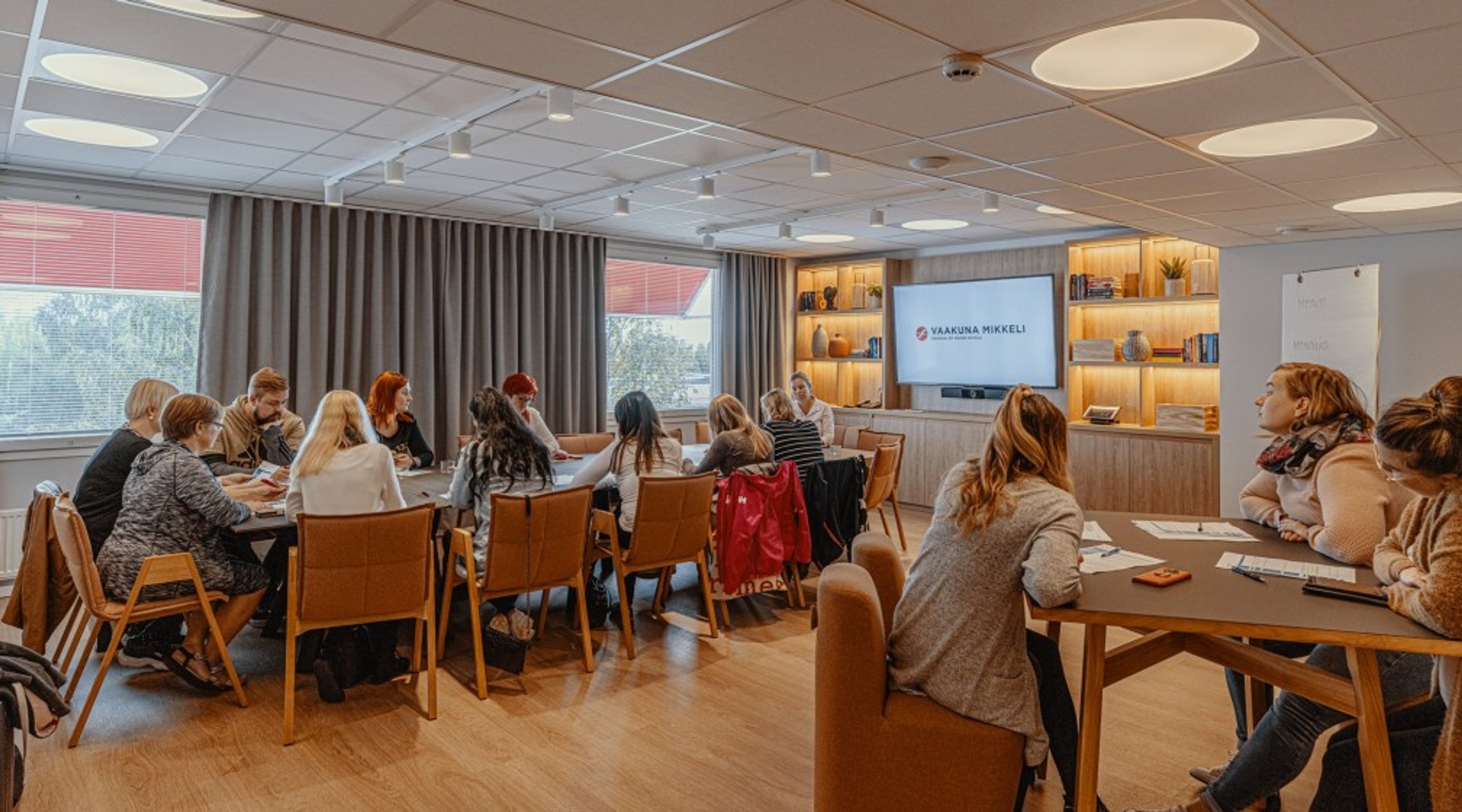 Valoisa kokoushuone, jossa ihmiset istuvat pöytien ääressä. Tilassa on puulattia, näyttö seinällä ja suuret ikkunat harmailla verhoilla.