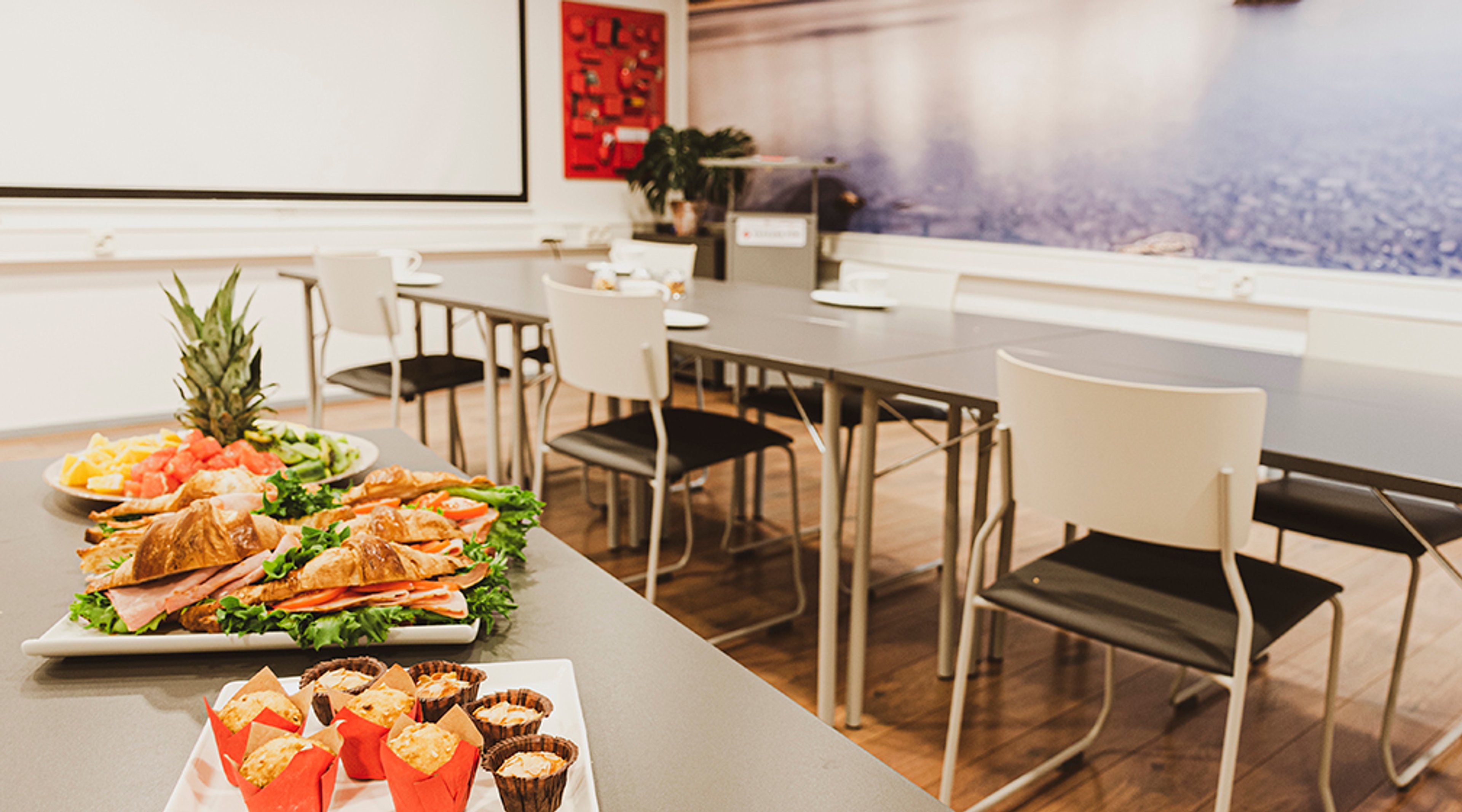Kokoustila, jossa valkoinen pöytä tuoleineen ja tarjoilupöytä täynnä voileipiä, hedelmiä ja leivonnaisia. Puinen lattia ja valoisat tilat.