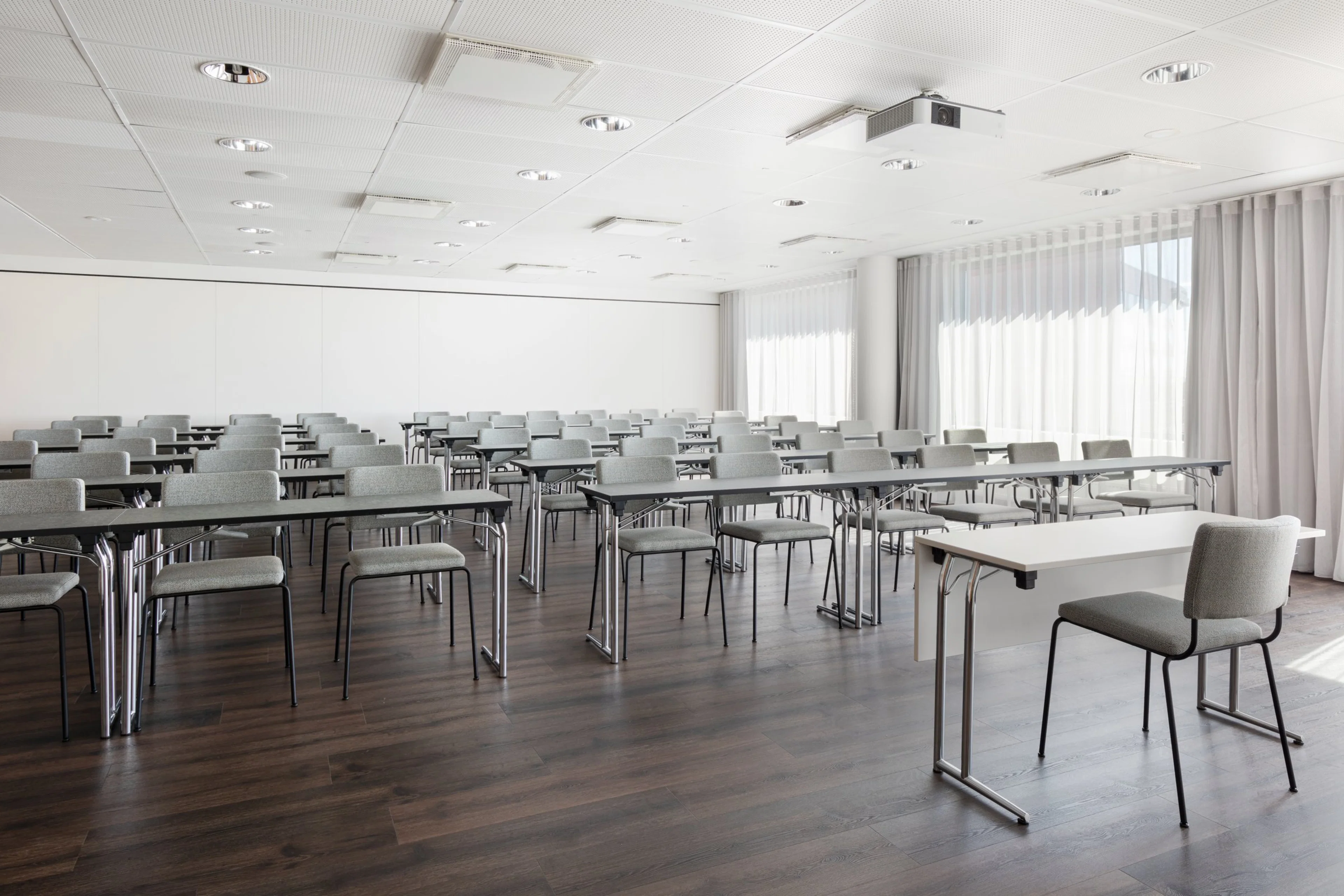 Valoisa seminaaritila valkoisilla seinillä, puulattialla ja harmailla tuoleilla. Kattoprojektori ja isot ikkunat vaaleilla verhoilla.