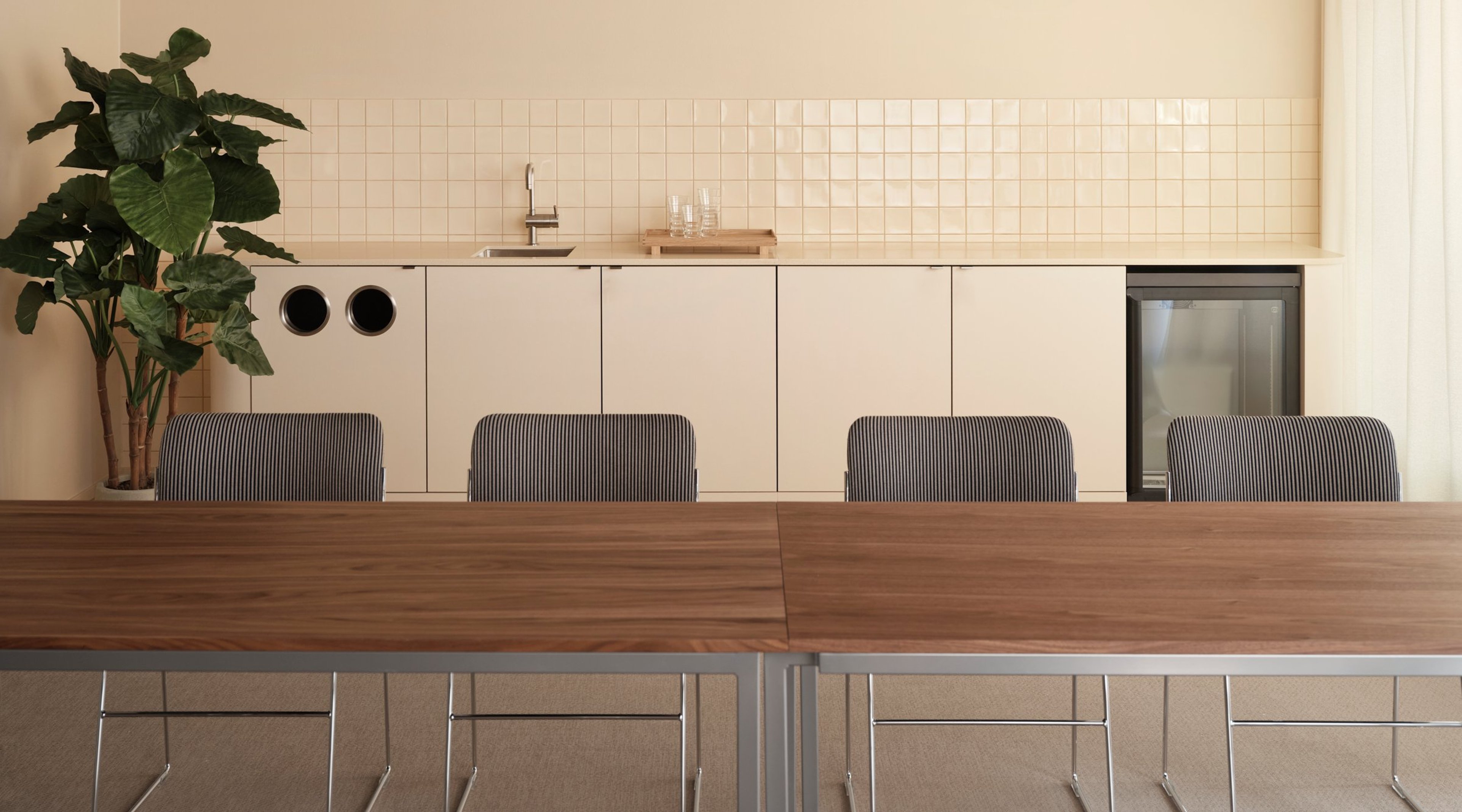 Minimalistinen neuvottelutila, jossa puinen pöytä, raidallisilla tuoleilla ja beige minikeittiö taustalla. Viherkasvit tuovat tilaan raikkautta.
