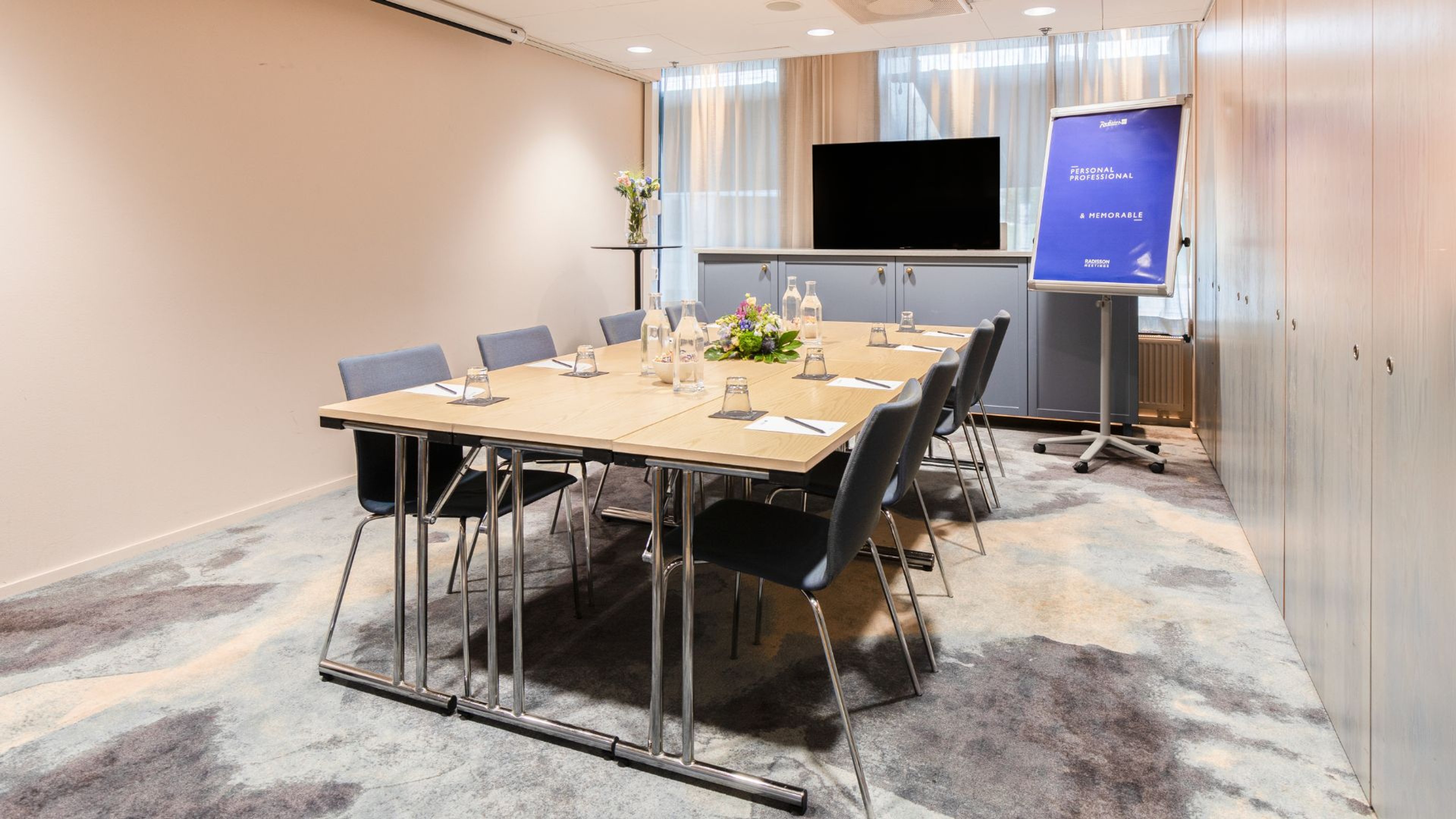 Moderni neuvotteluhuone puisella pöydällä, sinisillä tuoleilla, televisiolla ja esitysnäytöllä. Tilassa on tasainen kokolattiamatto ja hyvä valaistus.
