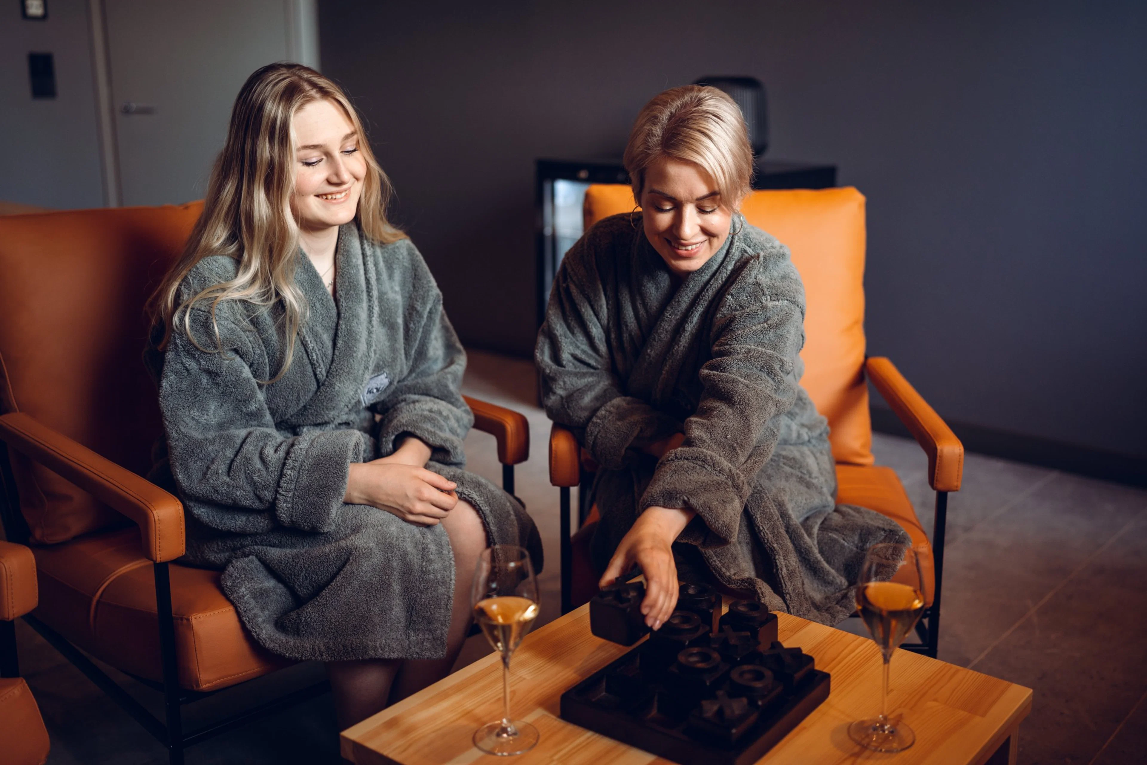 Kaksi henkilöä harmaissa kylpytakeissa istuvat oransseissa tuoleissa puisen pöydän äärellä, jolla on laseja ja tarjotin.