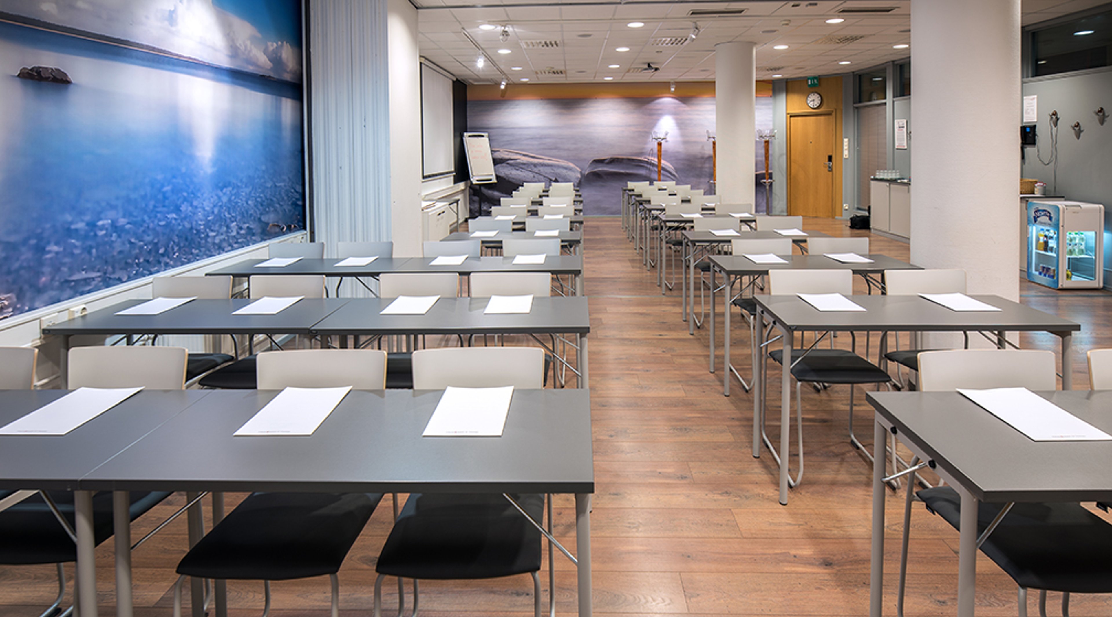Kokoustila luokkamuodossa, jossa pöydät rivissä. Seinillä sininen merimaisematapetti. Puulattia ja valkoinen katto valaistuksella.