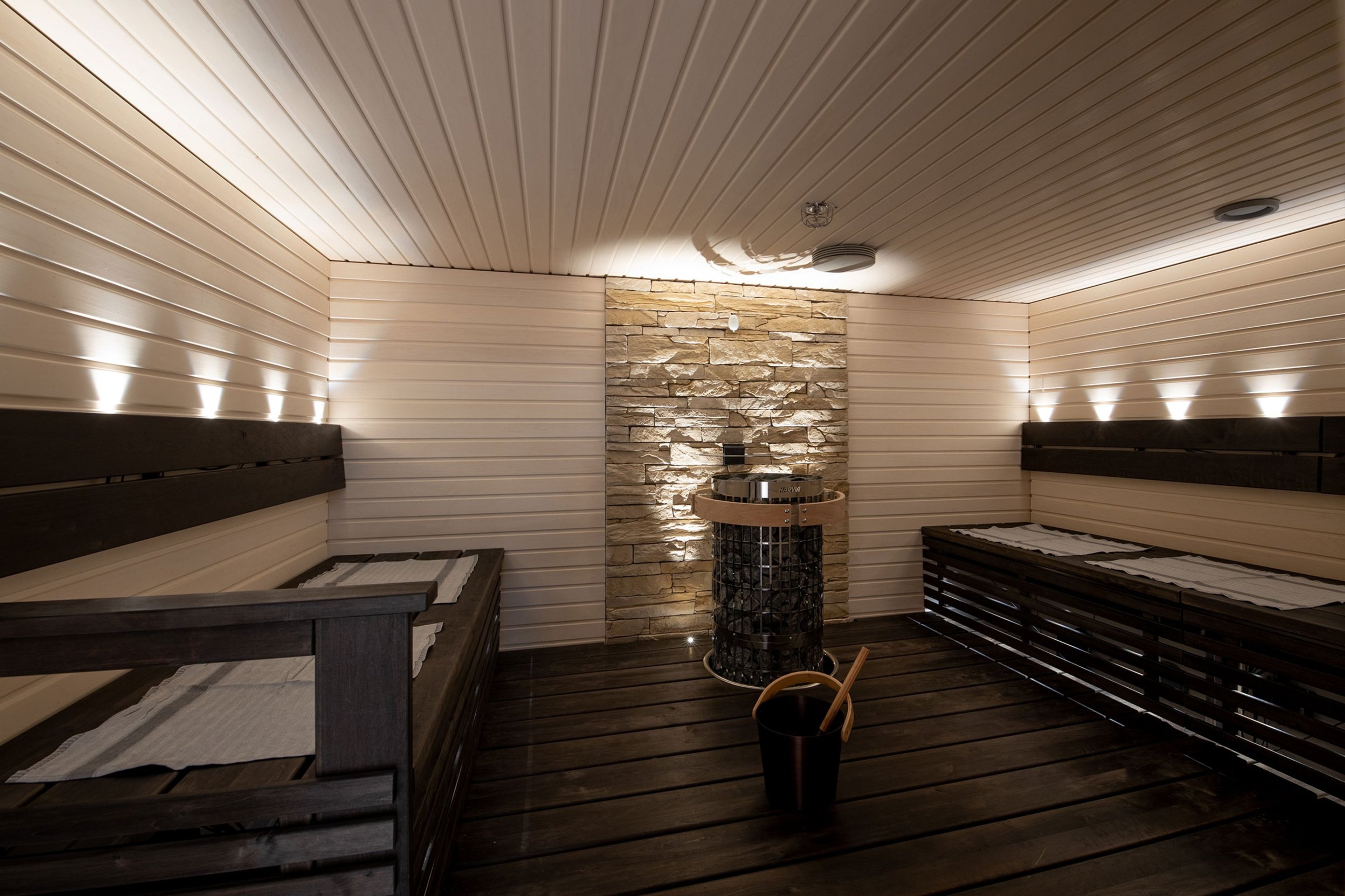 Moderni sauna valkoisilla paneeliseinillä, kivisellä takaseinällä ja kiukaalla keskellä. Tummat lauteet molemmin puolin.