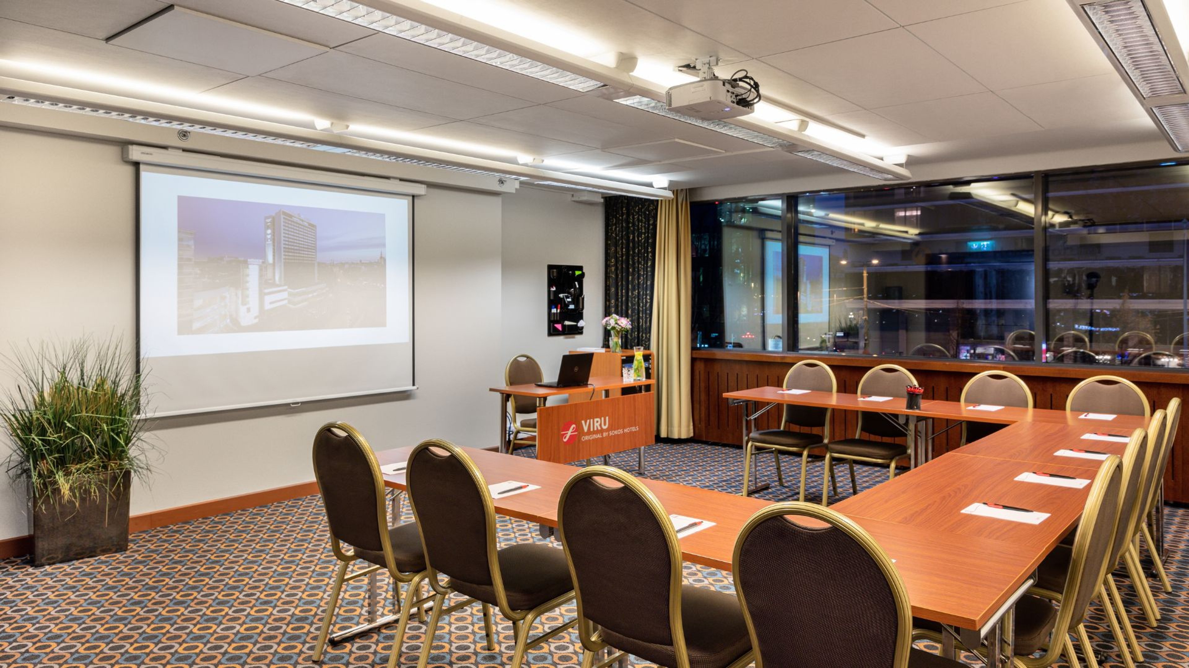 Kokoushuone puisilla pöydillä ja tuoleilla, projektorikangas seinällä, tasainen kokolattiamatto ja hyvä valaistus.