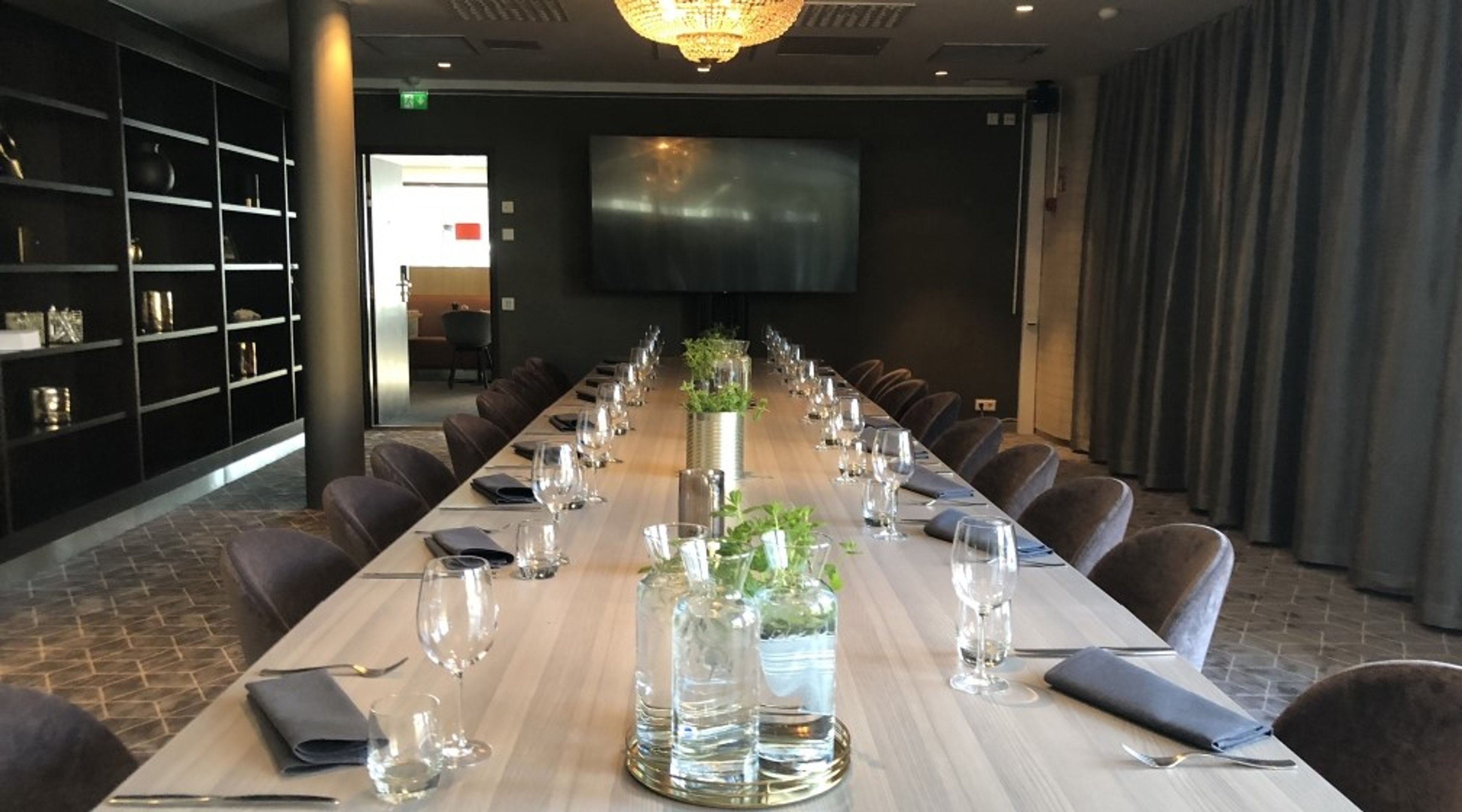 Pitkä neuvottelupöytä, jossa ruskeita tuoleja, laseja ja viherkasveja keskellä. Taustalla näyttö, kristallikruunu katossa ja hyllykkö.